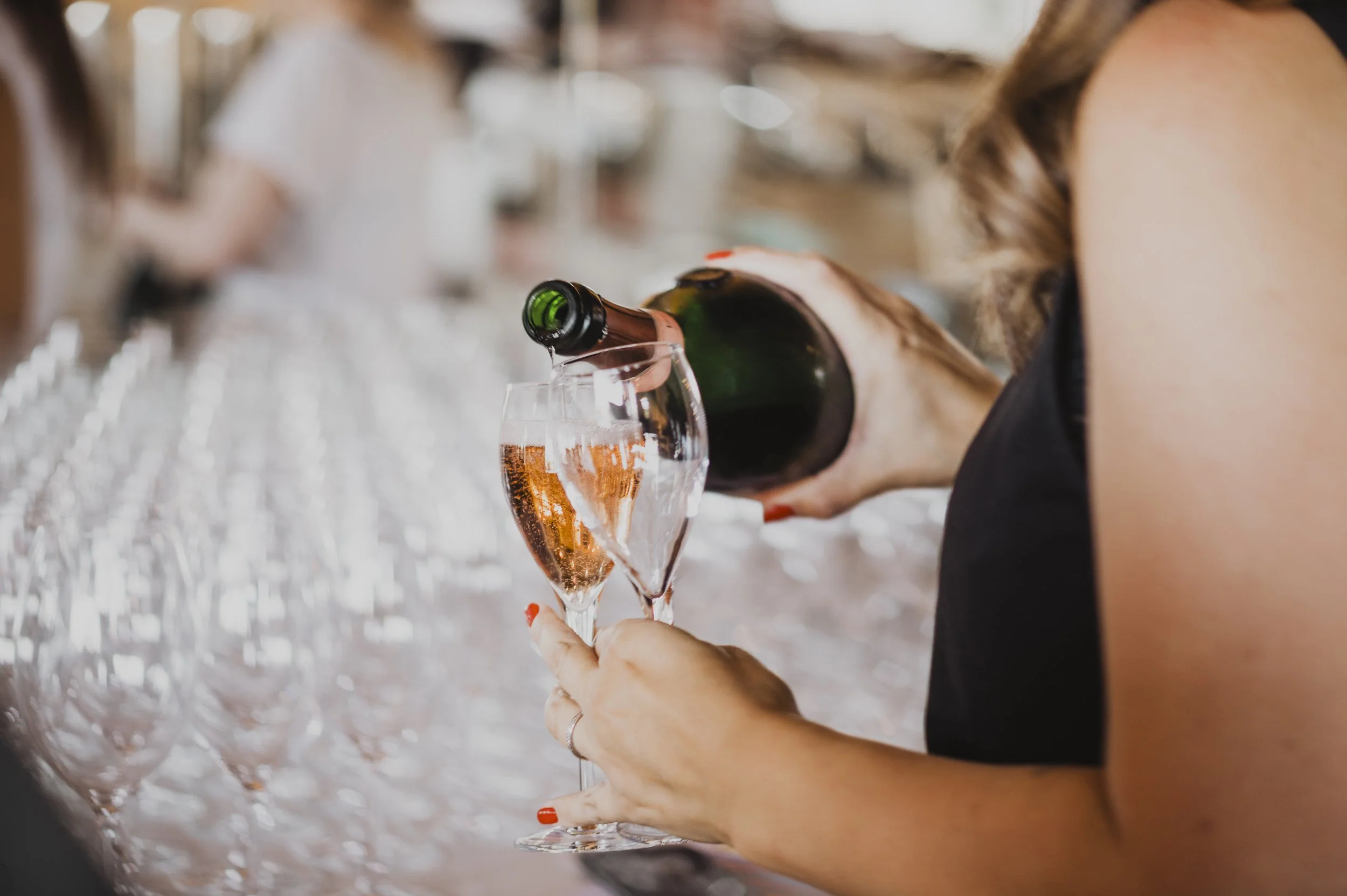 A woman pouring rosé wine into a wine glass at a bar or event with many empty glasses in the background.