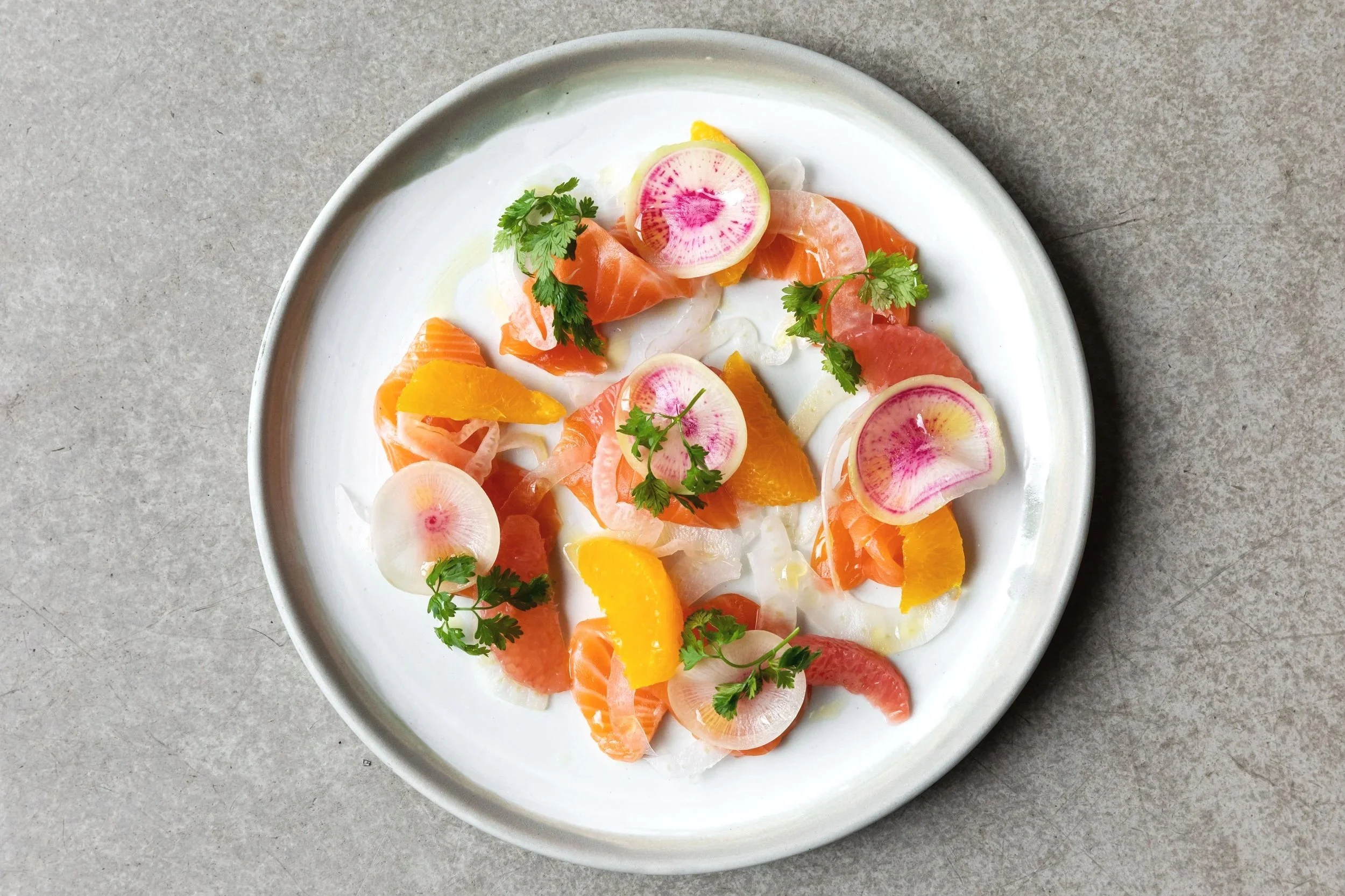 A white plate containing smoked salmon, thinly sliced radishes with pink centers, orange segments, and fresh green cilantro leaves, placed on a grey surface.