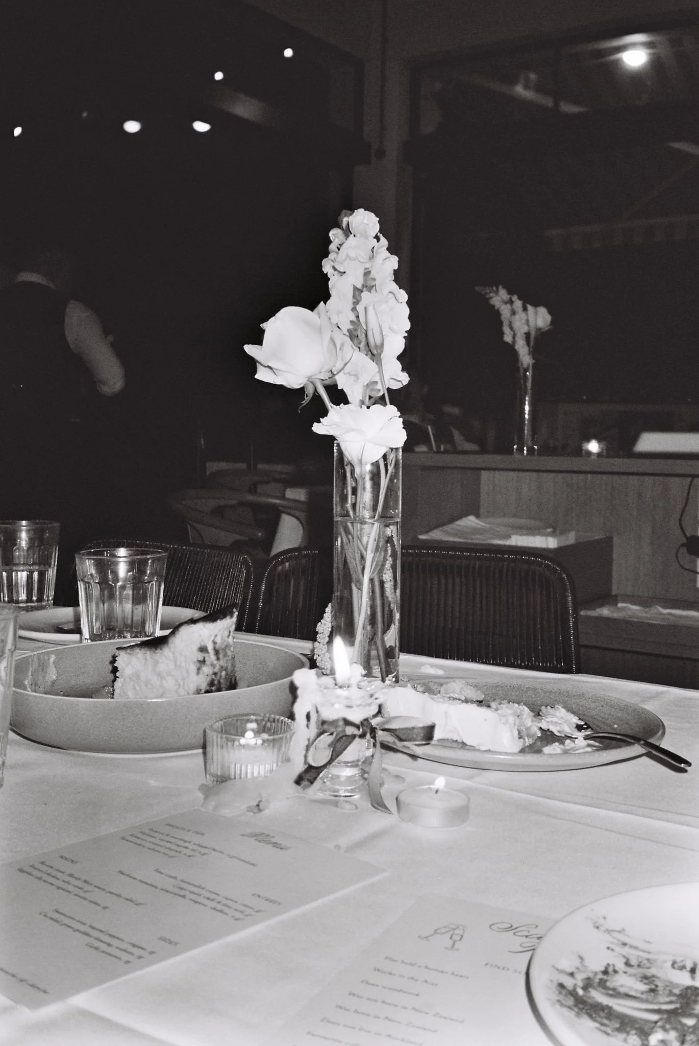 A table setting with a vase of flowers and a cake slice on a plate in a dimly lit restaurant. There are glasses of water and a menu on the table.