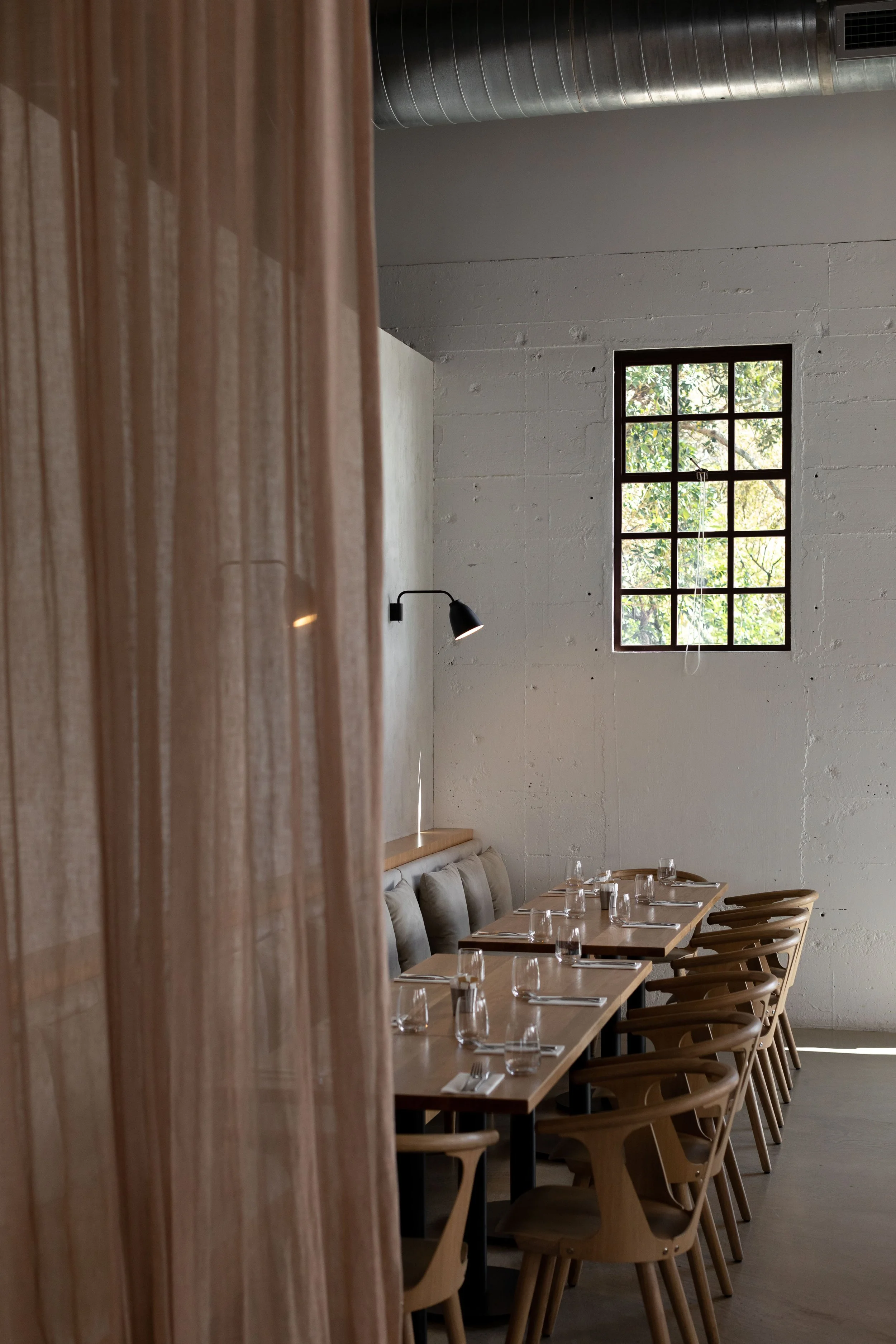 Interior of a minimalist restaurant or cafe with a long wooden table set with glasses and napkins, surrounded by matching wooden chairs, against a white wall with a window showing greenery outside, and natural light illuminating the space.