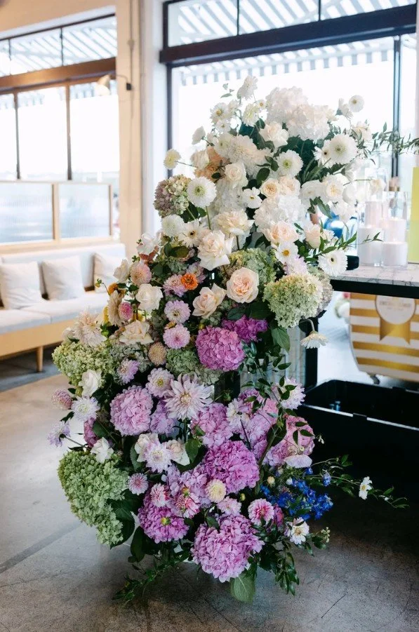 Large wedding floral arrangement with white, light pink, purple, and green flowers in an indoor setting near large windows with seating area.