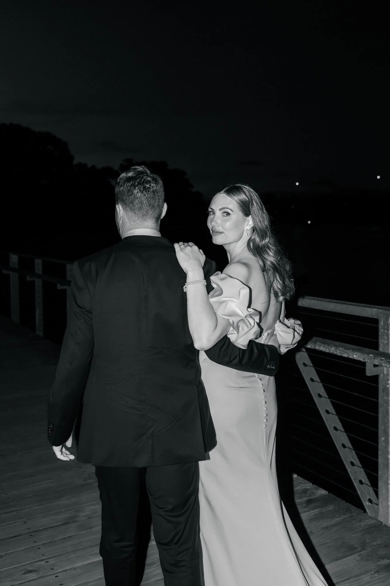 A woman in a wedding dress with ruffled sleeves and pearl bracelet, facing the camera, holding hands with a man in a black tuxedo, outdoors at night with a dark sky and horizon in the background.