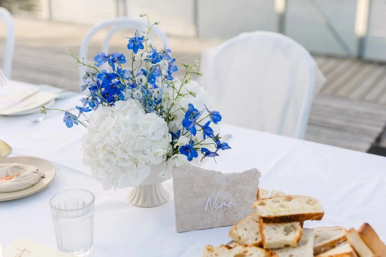A wedding table set with a white tablecloth, featuring a bouquet of white hydrangeas and blue delphiniums in a white vase, a marble table card labeled 'Nina', a glass of water, a plate of sliced bread, and some linens in the background.
