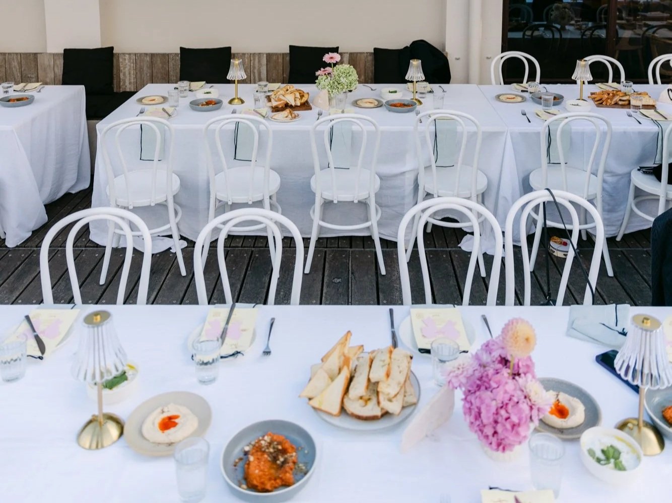 Wedding banquet table setting with white tablecloth, white chairs, floral centerpiece, plates of food, and small lamps.