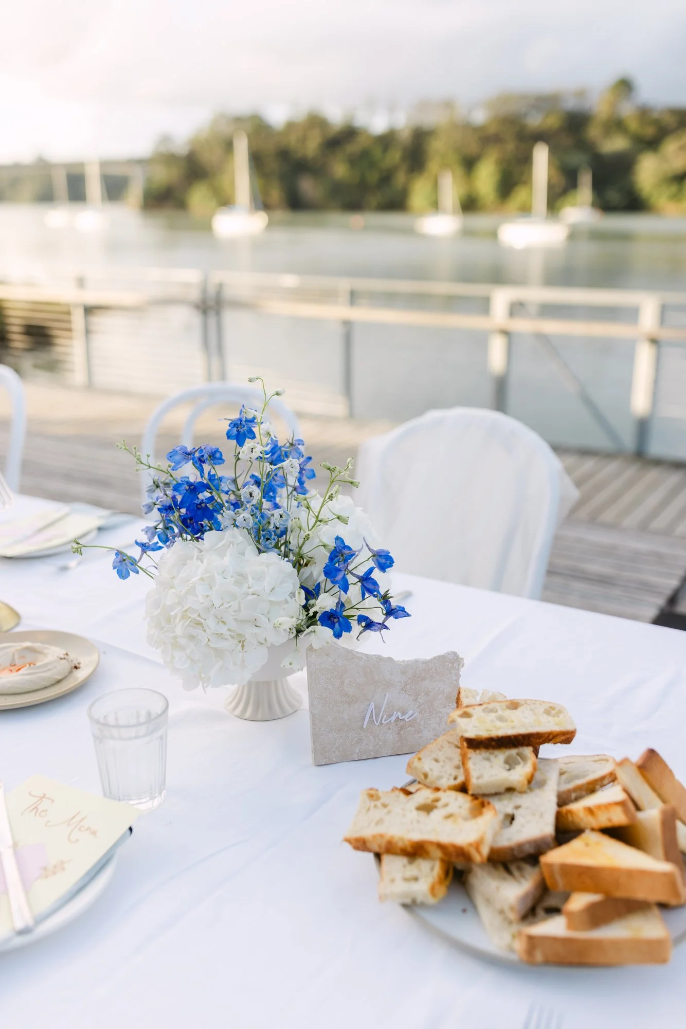 A wedding table with a bouquet of blue and white flowers, a small card with the name 'Nina,' some sliced bread, and a menu at an outdoor event by a body of water with sailboats in the background.
