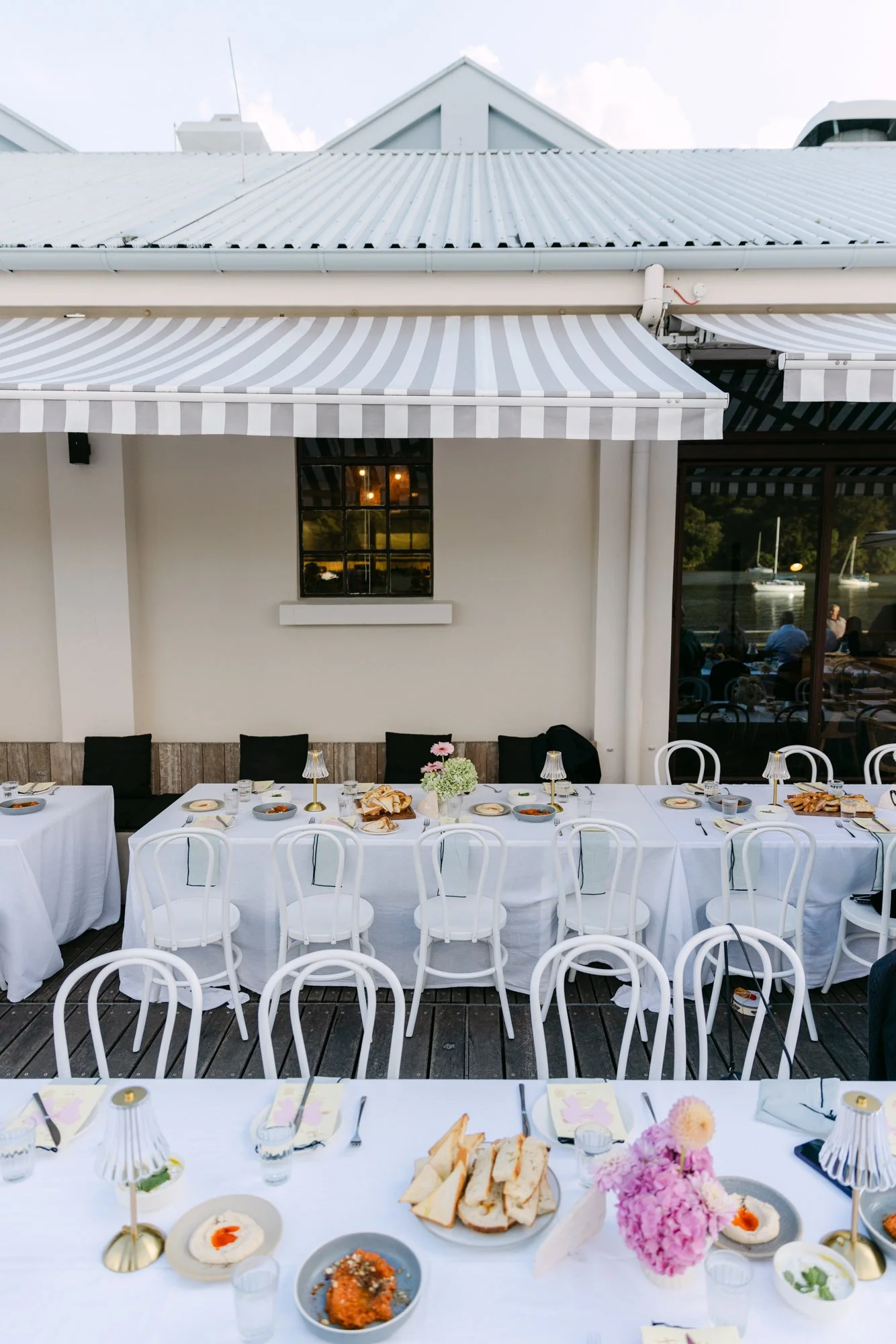 Outdoor dining setup with a long table covered in white tablecloth, decorated with pink flowers, surrounded by white chairs. Plates with food, bread, and small desserts are on the table. The background features a building with white walls, large wind