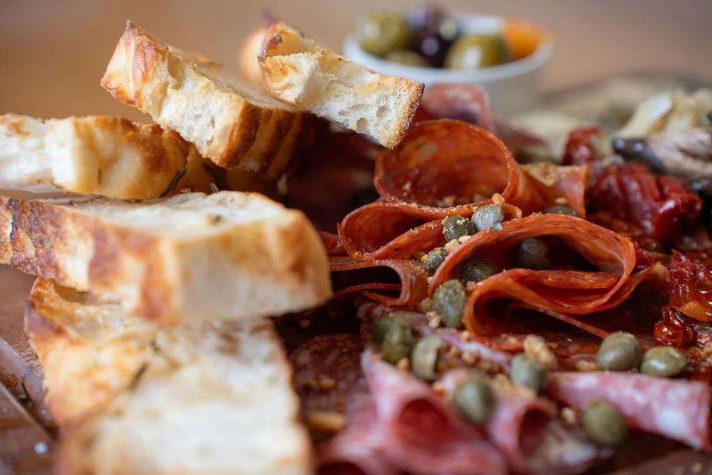 Close-up of a charcuterie board with slices of toasted bread, cured meats like salami and ham, and capers, with a small bowl of olives in the background.