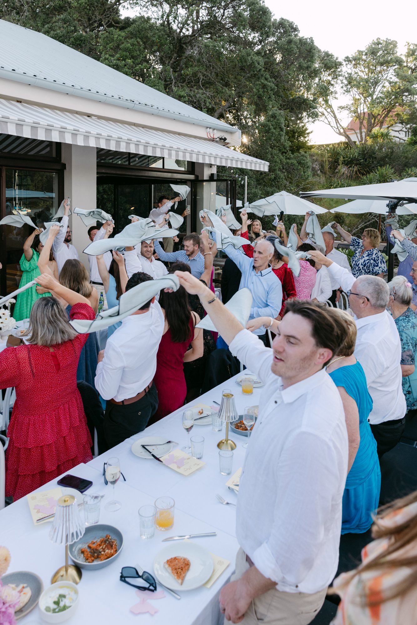 A group of people at a wedding or gathering outdoors, dancing and waving cloth napkins in the air around a long table with food and drinks, under umbrellas and near a house with trees in the background.