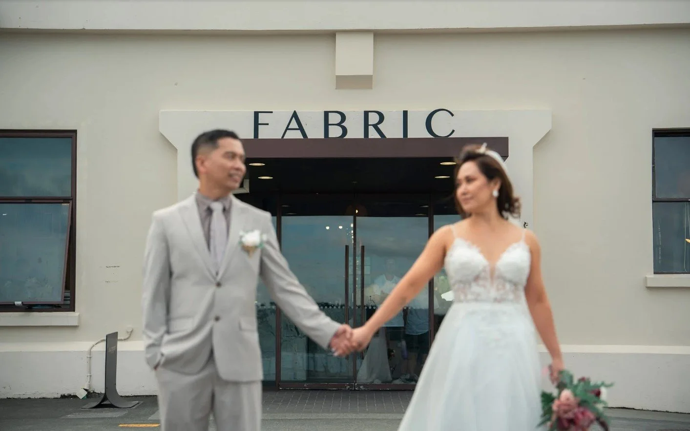 A bride and groom holding hands and smiling outside a building marked 'FABRIC' during a wedding ceremony.