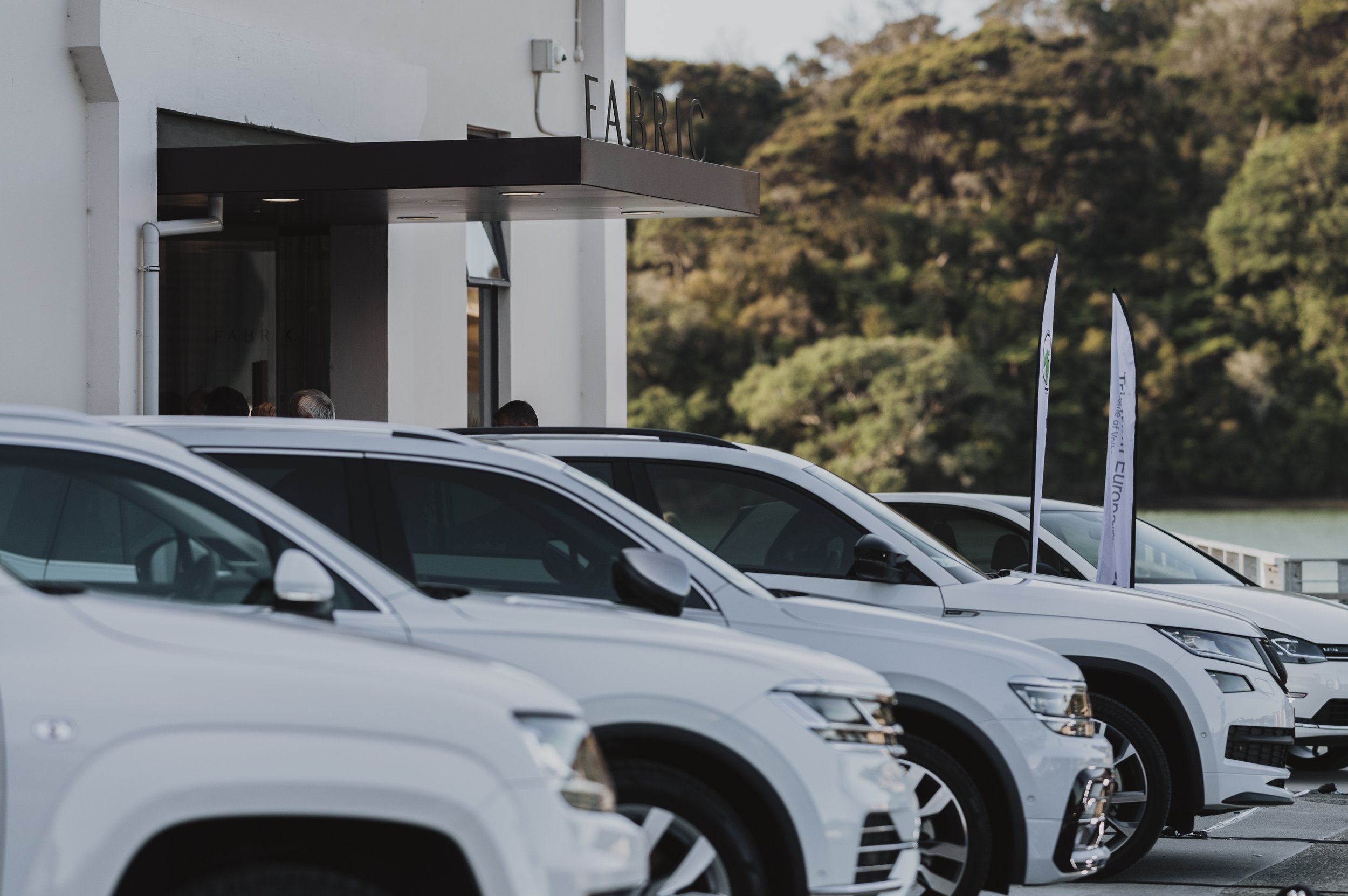 Line of white cars parked outside a building with a sign that reads 'FABRIC,' in front of a hillside with trees.
