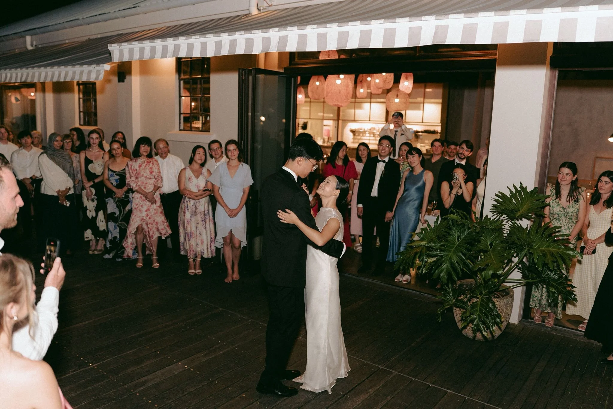 Couple dancing at a wedding reception with a group of people watching inside a warmly lit venue.