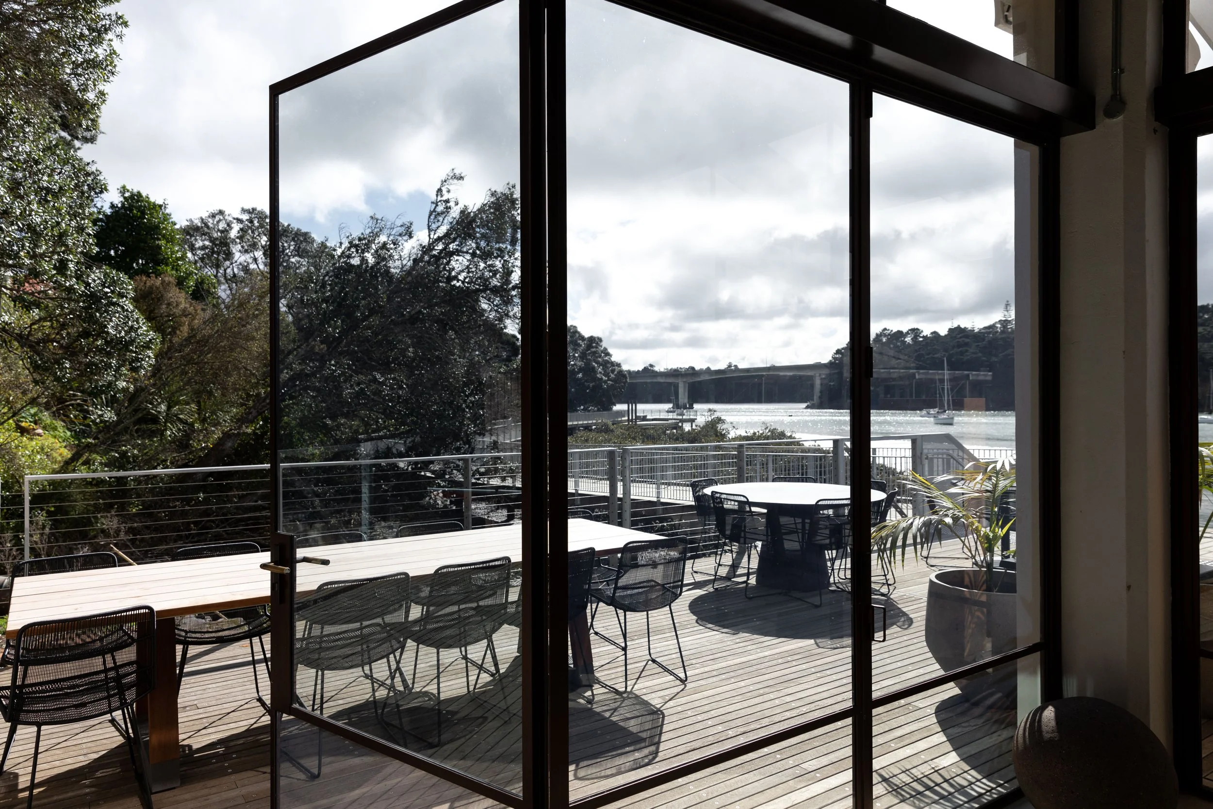 View of an outdoor balcony with black metal chairs and a wooden dining table, overlooking the harbour, trees, and a bridge under a cloudy sky, seen through glass sliding doors.