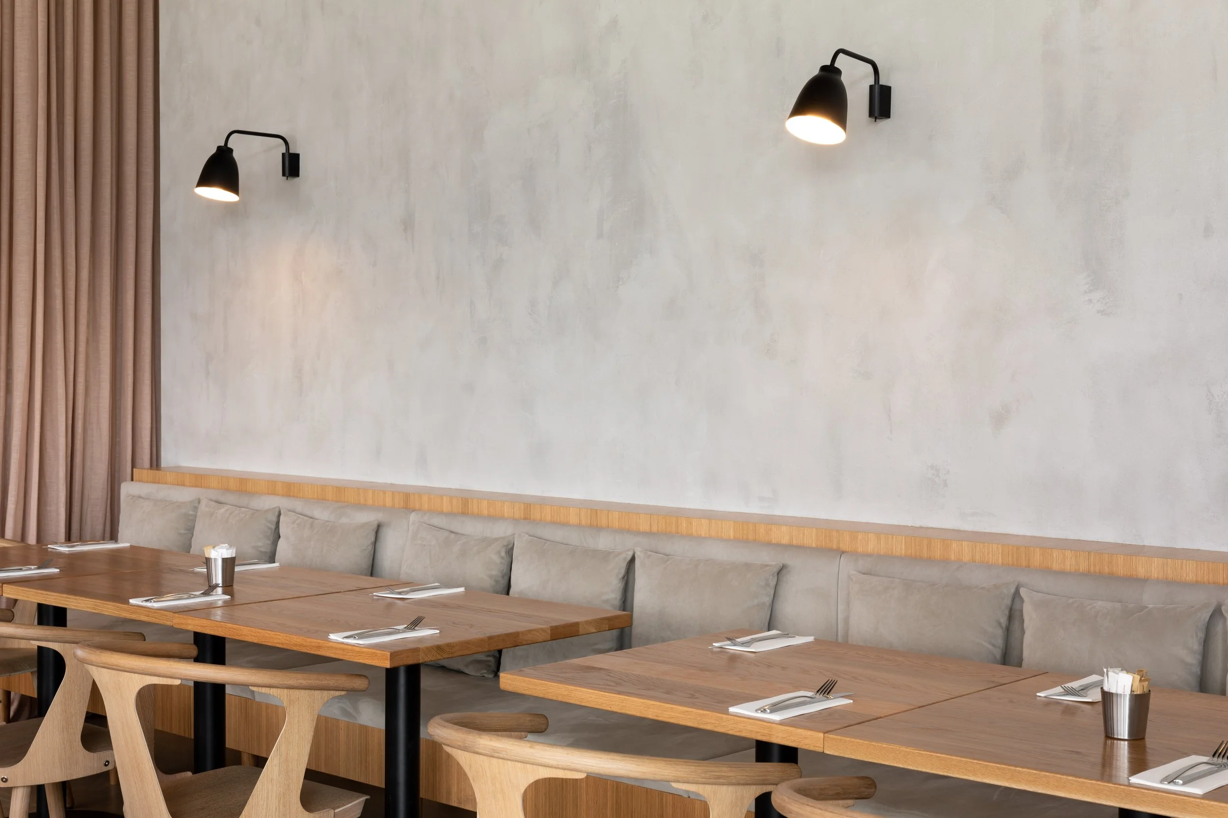 Empty restaurant dining area with wooden tables, light wood chairs, beige cushioned bench, and black wall-mounted lamps on a textured gray wall.