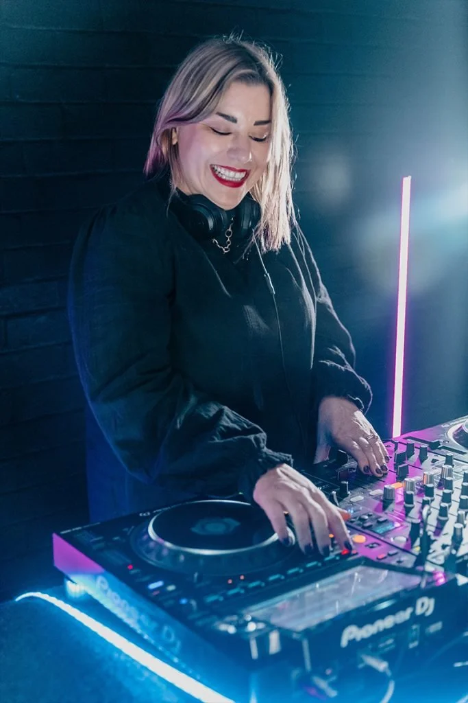 A woman DJ with blonde hair, wearing a black jacket, smiling while operating a Pioneer DJ controller with colorful lights.