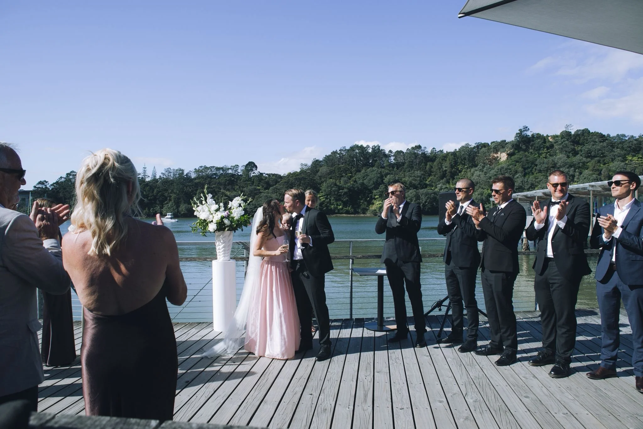 A wedding ceremony taking place outdoors on a wooden deck by the water, with a bride and groom kissing surrounded by guests dressed in formal attire, against a backdrop of trees and a blue sky.