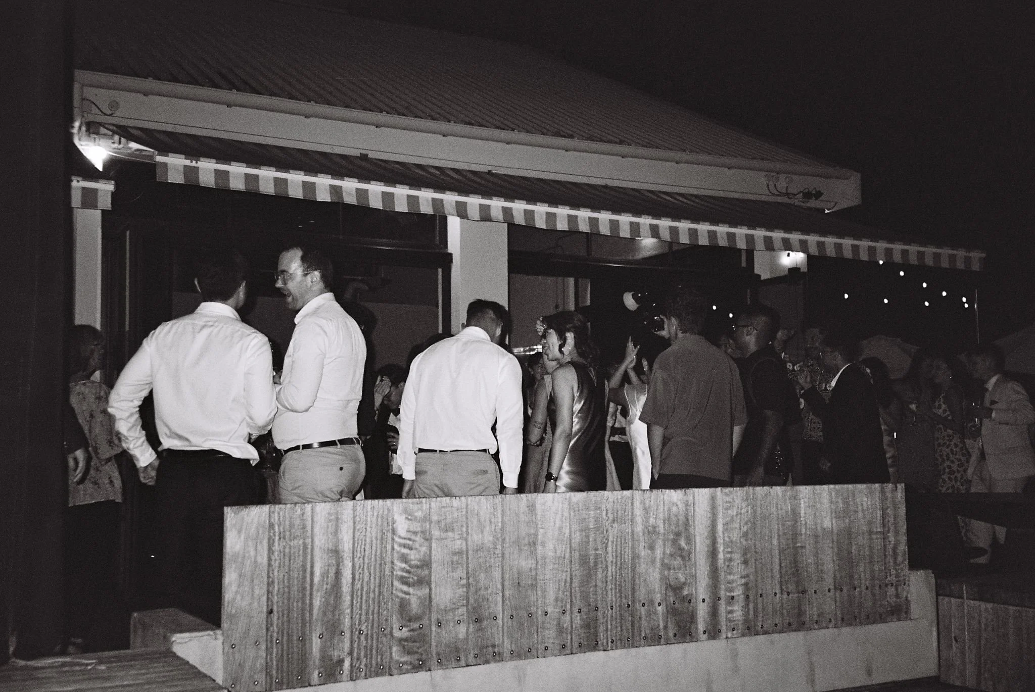 People gathered at an outdoor event at night, engaged in conversations, under a striped awning with a building in the background.