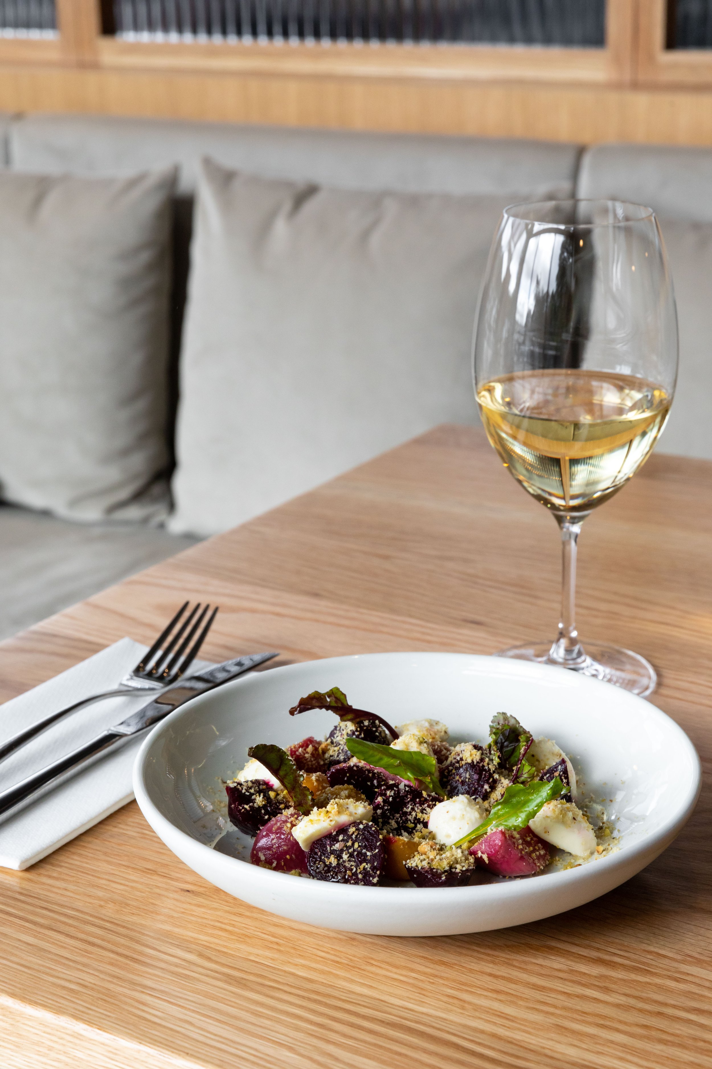 Glass of white wine on a wooden table with a plate of salad and cutlery in a restaurant setting.
