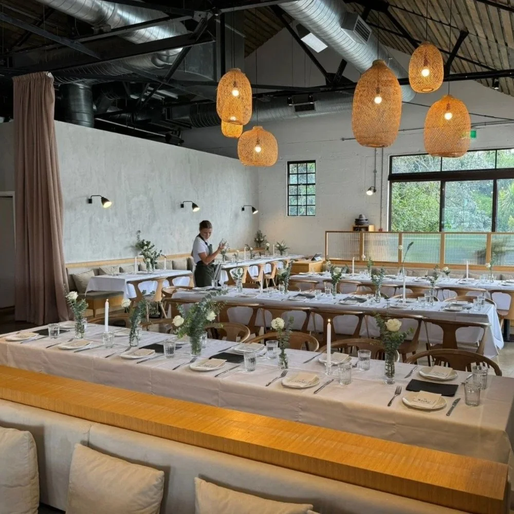 Interior of a decorated restaurant or wedding space with arranged tables, hanging woven pendant lights, and a person preparing the table.