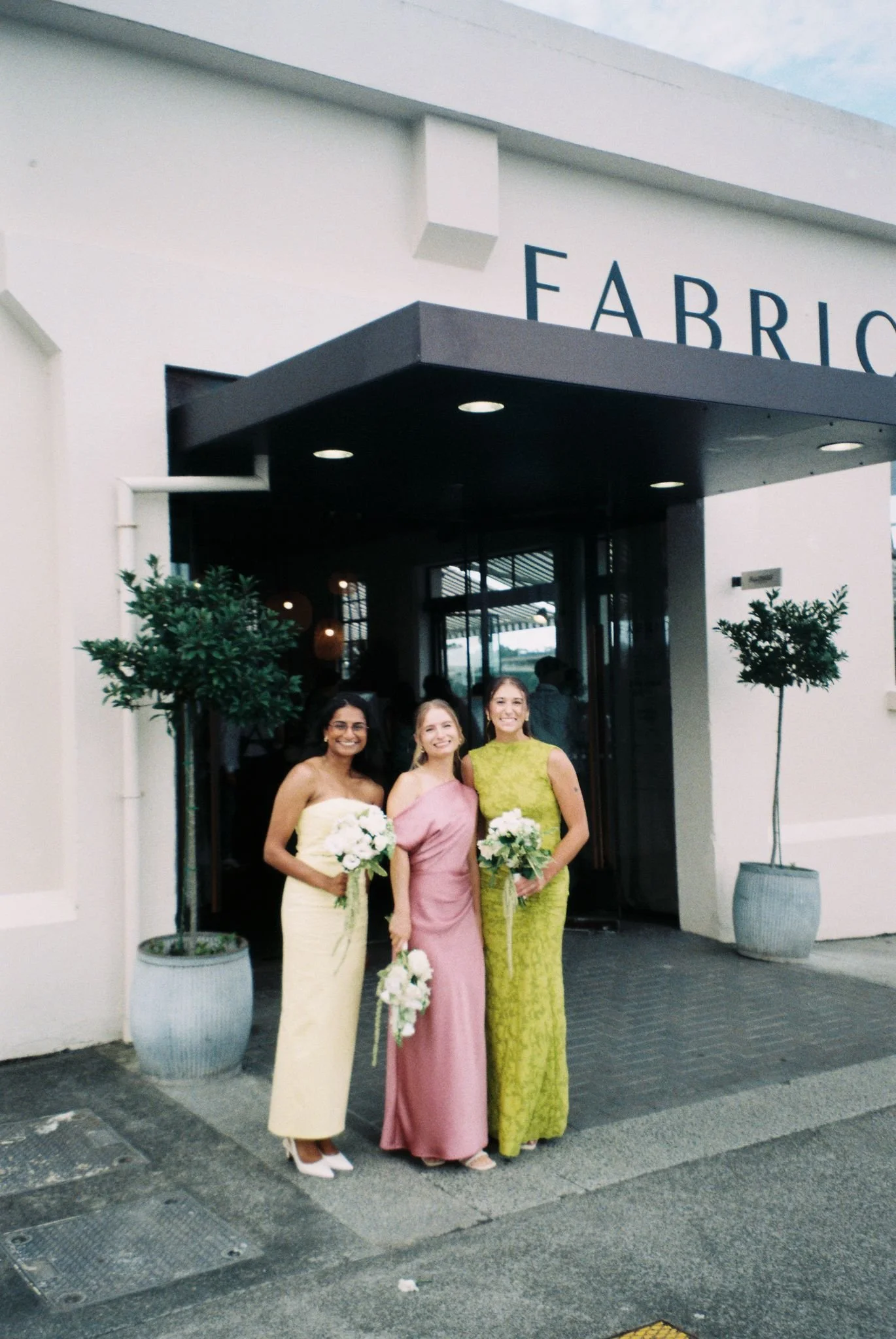 Three women wearing elegant dresses holding bouquets of white flowers, standing outside a modern building with the sign 'Fabrik' above the entrance. Two potted plants are on either side of the entrance, and a few people are visible inside through the