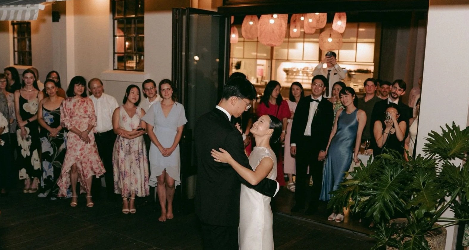 A couple dancing closely at a wedding reception, with guests watching and taking photos in a warmly lit indoor setting.