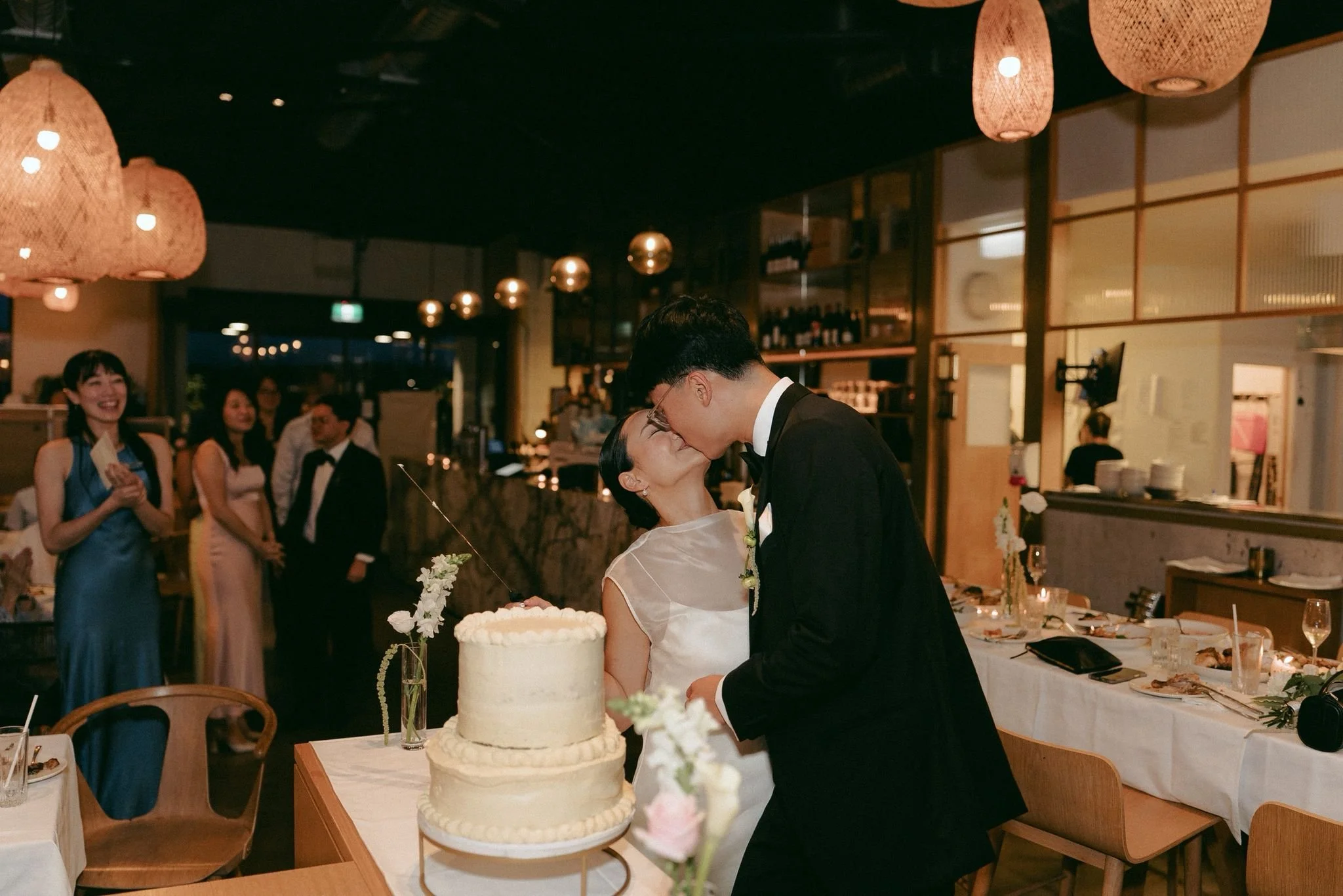 A newlywed couple is kissing at their wedding reception, with a wedding cake in the foreground and guests smiling in the background.