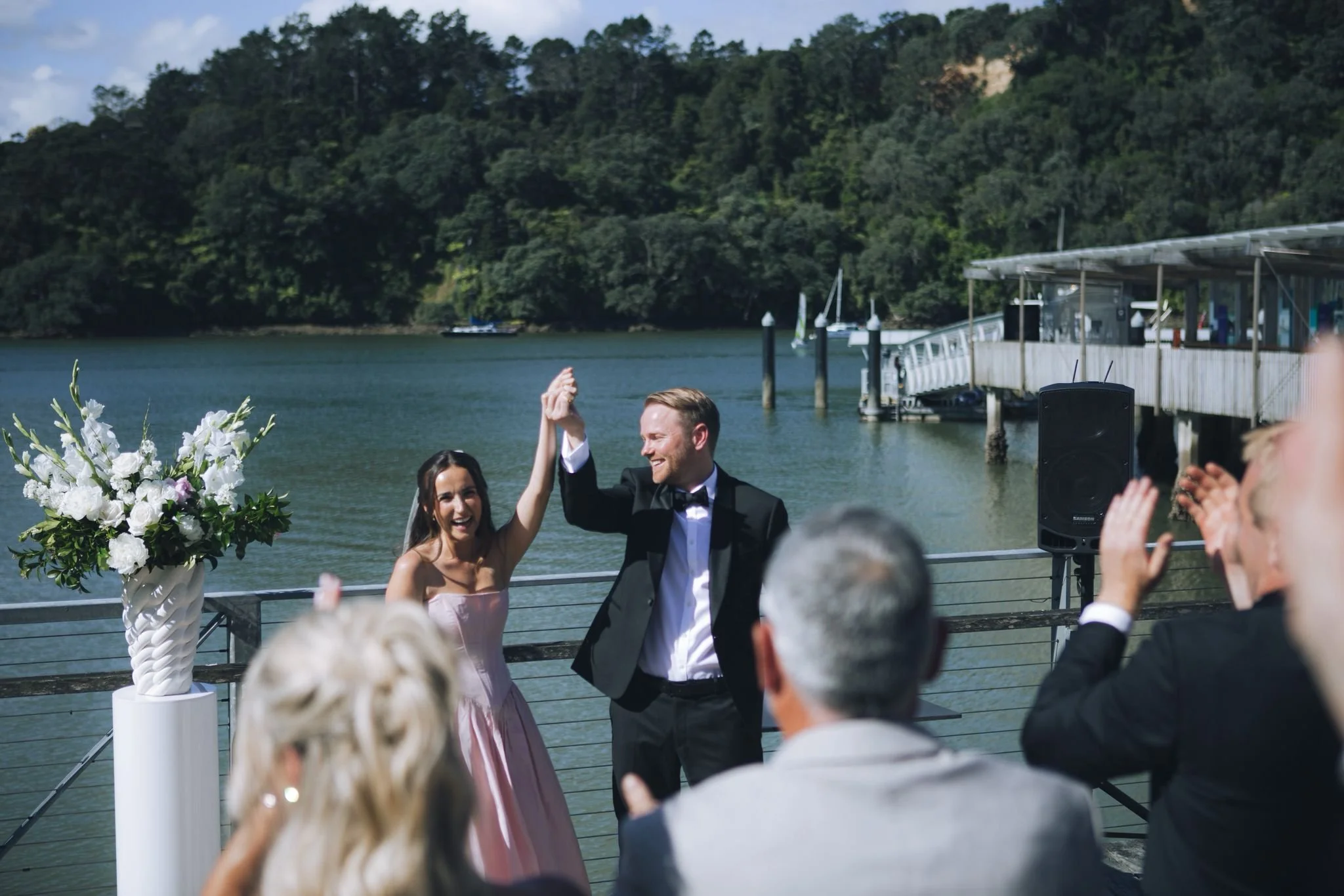 A newlywed couple, the bride in a pink dress and the groom in a black tuxedo, dancing and smiling at an outdoor wedding reception by the water with guests watching, trees, sailboats, and a pier in the background.