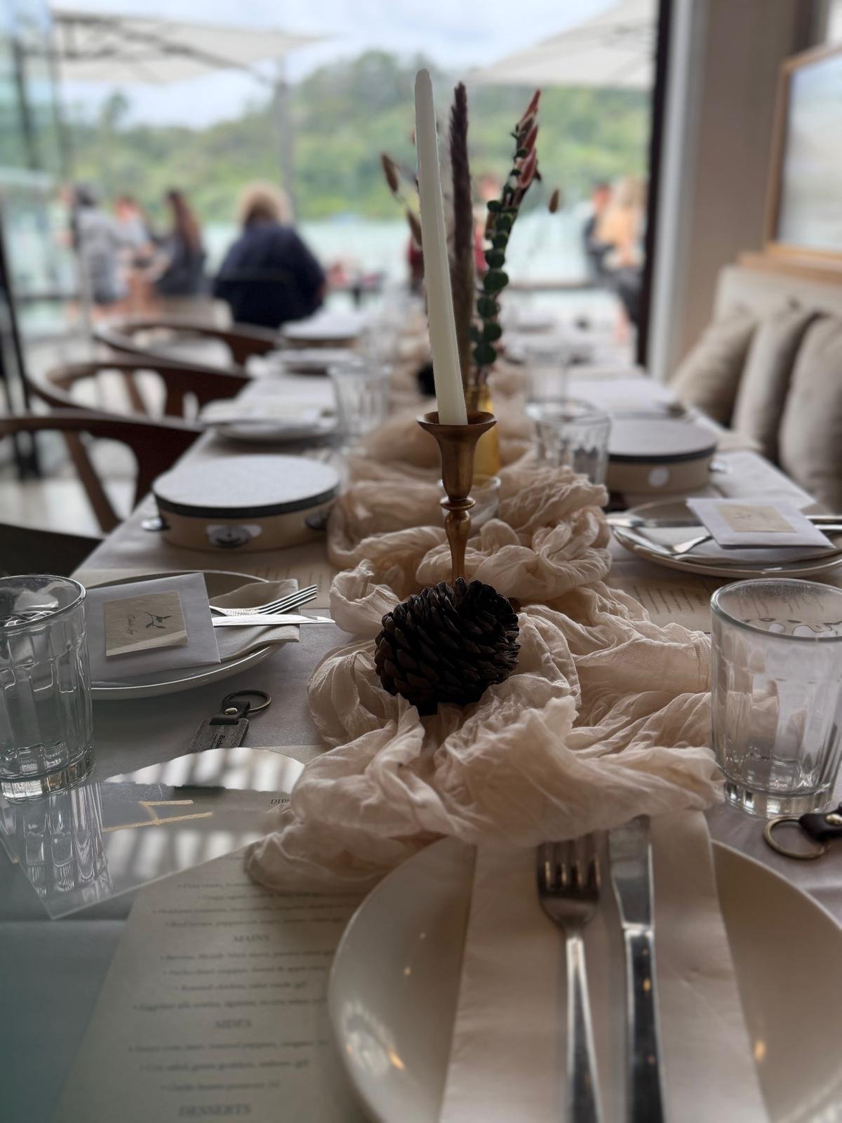 A dining table set for a meal with plates, silverware, glasses, and a centerpiece featuring a candle with a pinecone at its base, on a cream-colored fabric runner, in a restaurant with large windows and blurred people in the background.
