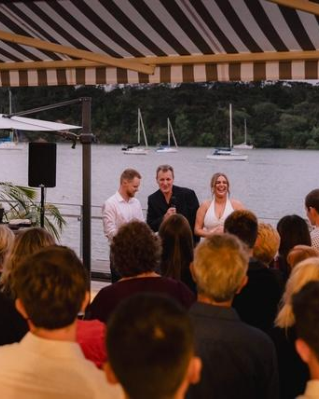 People attending an outdoor wedding by the water, with sailboats and trees in the background, under a striped canopy.
