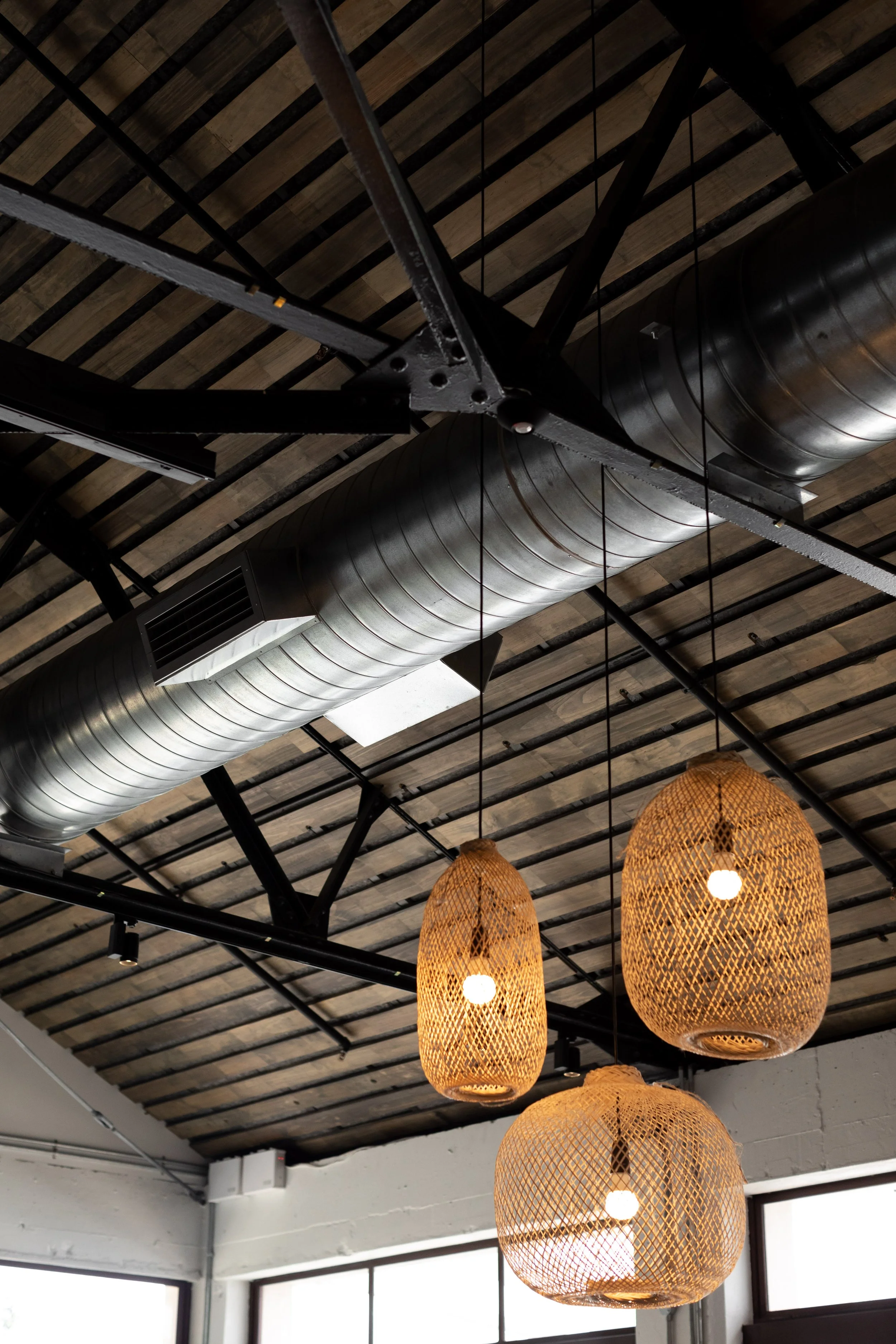 Interior ceiling with black metal beams, large cylindrical black air duct, and woven rattan pendant lights hanging at different heights.