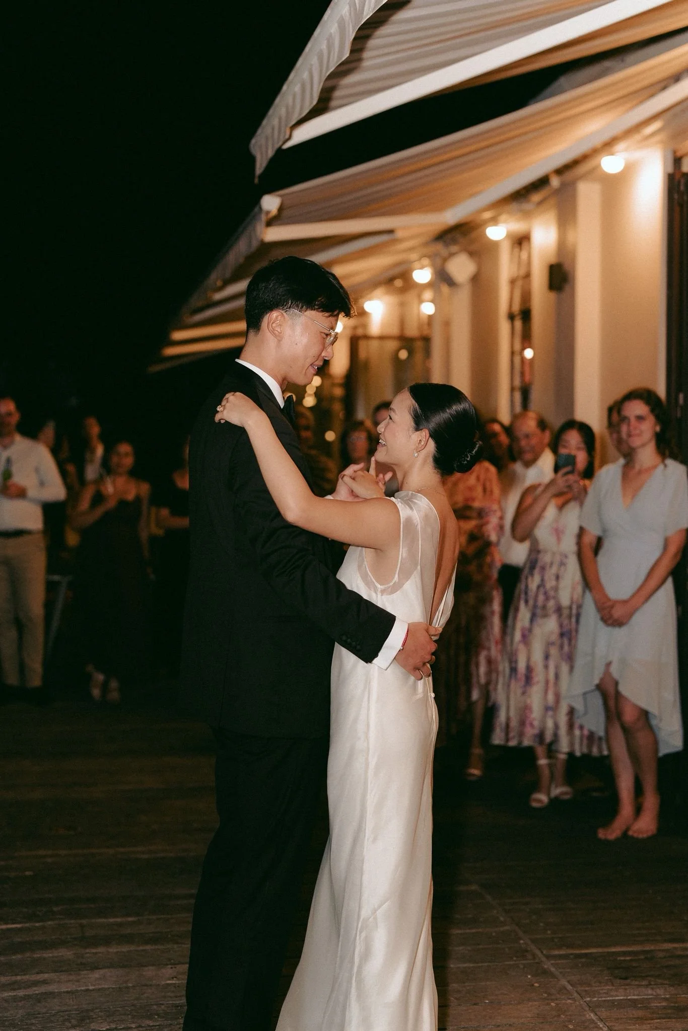 A man and woman dance closely at a wedding reception, with guests watching in the background under warm lighting.