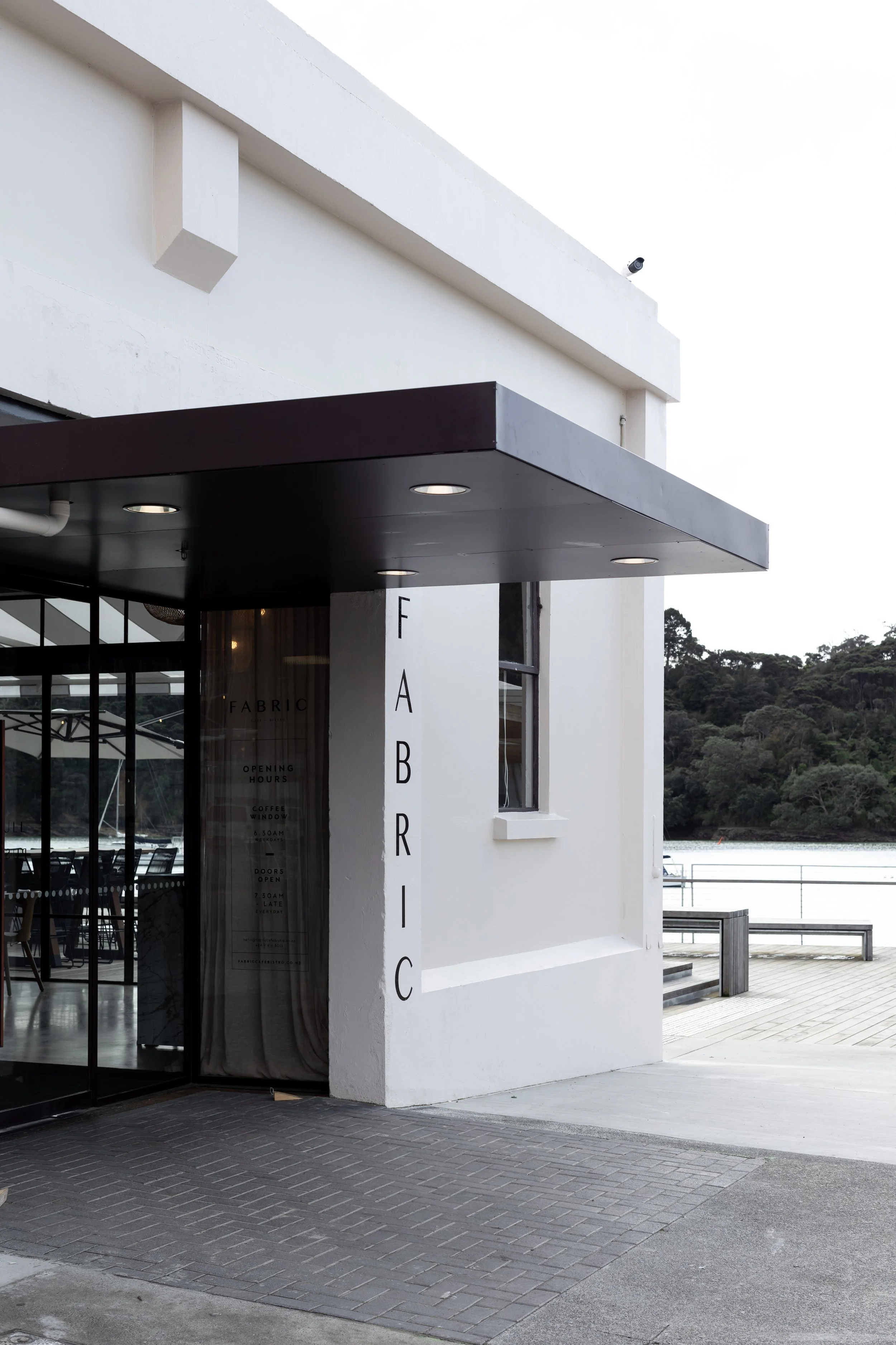 A modern white building with a dark overhang, large glass windows, and a sign that reads 'FABRIC'. There is a waterfront view with trees and water in the background, and part of a palm tree is visible to the side.