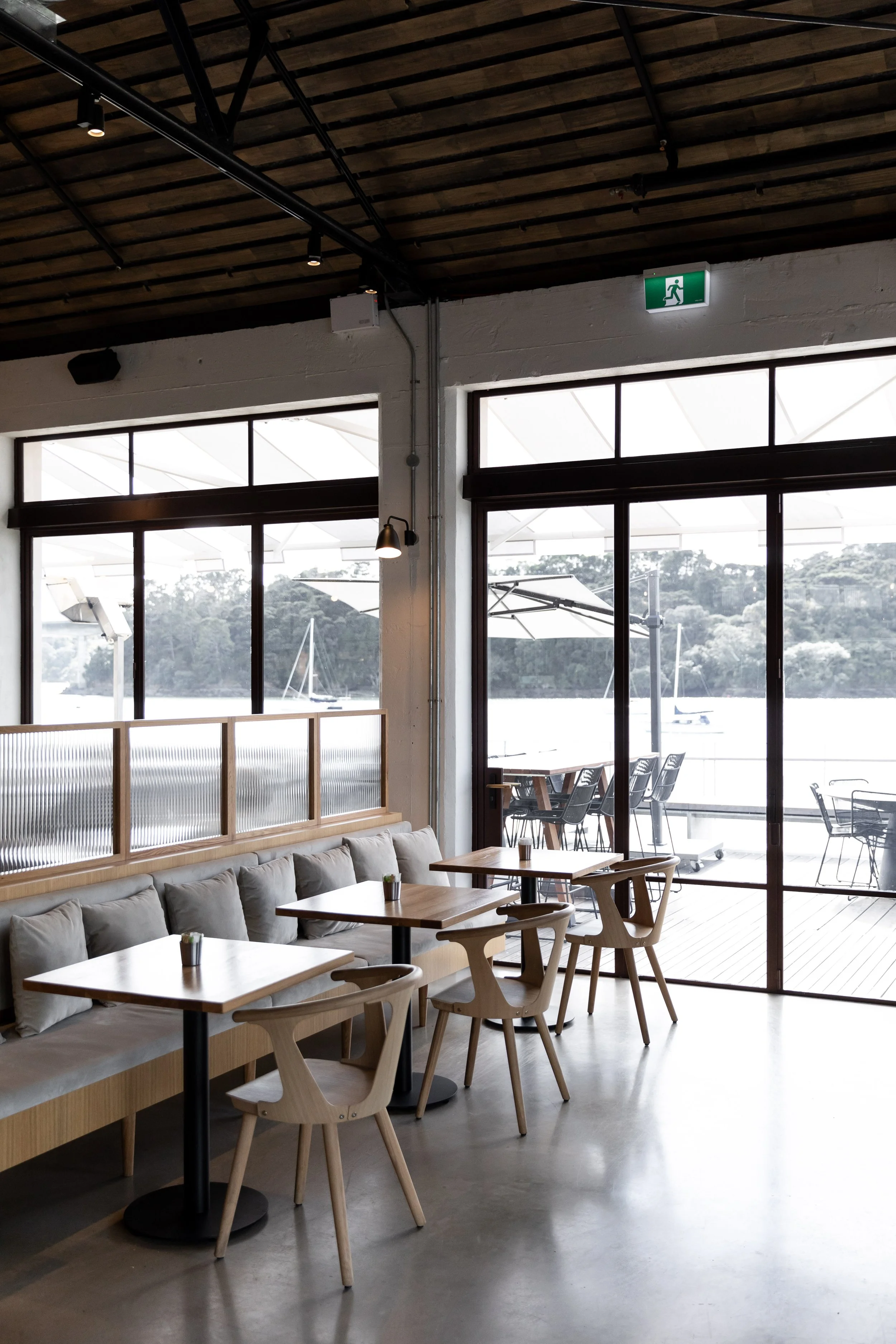 Interior of a modern cafe with wooden tables and chairs, large windows overlooking a river or lake with sailboats, and an emergency exit sign above the door.