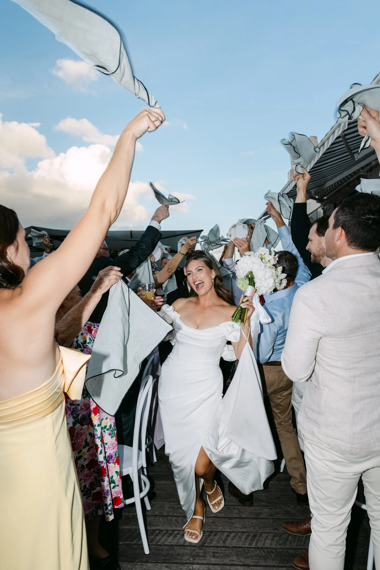 A group of people celebrating a wedding outdoors, on a deck or patio, with the bride in a white dress holding a bouquet of white flowers, smiling and surrounded by friends and family holding napkins or cloths in the air.
