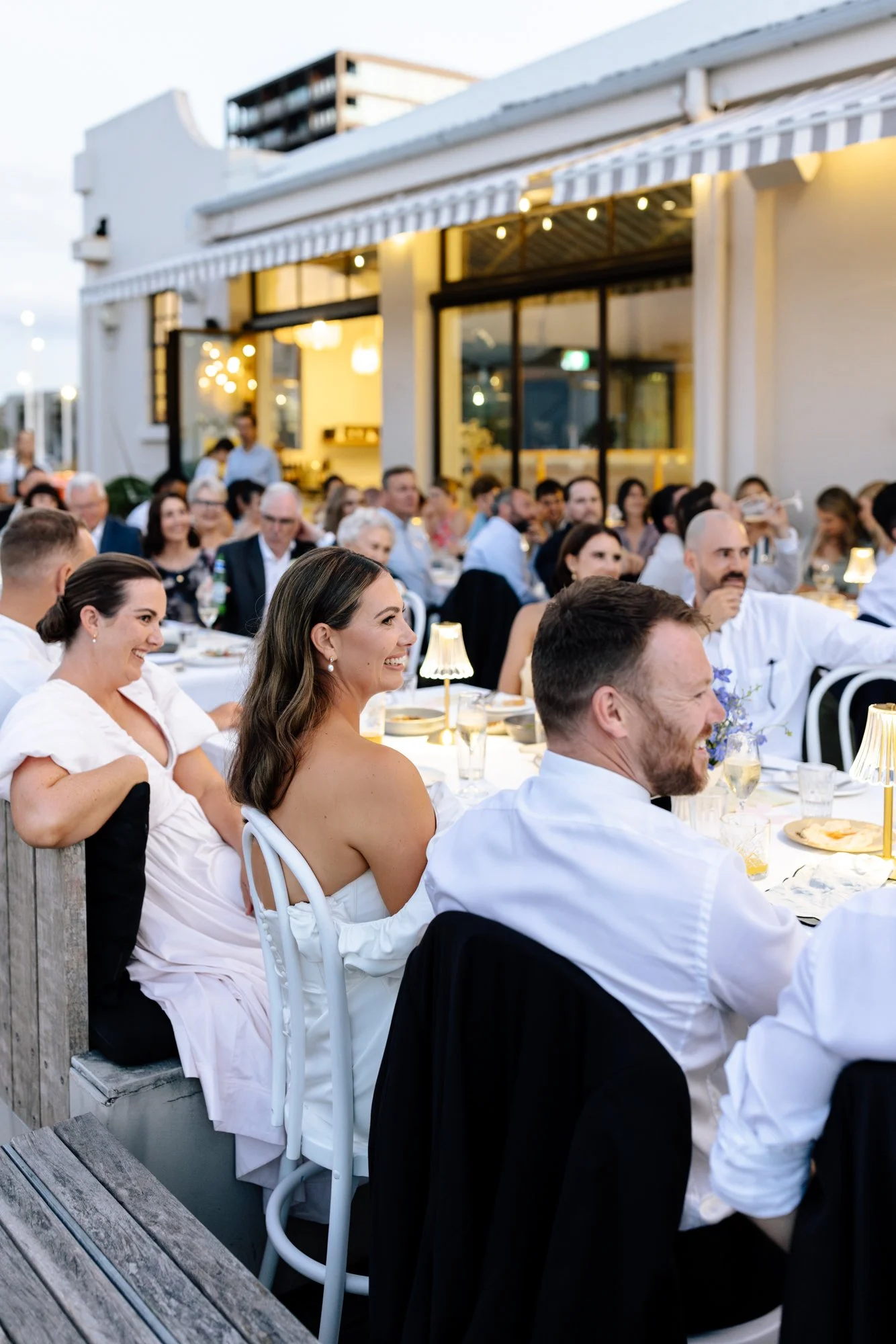 People seated at tables during an outdoor event with string lights and a building in the background.