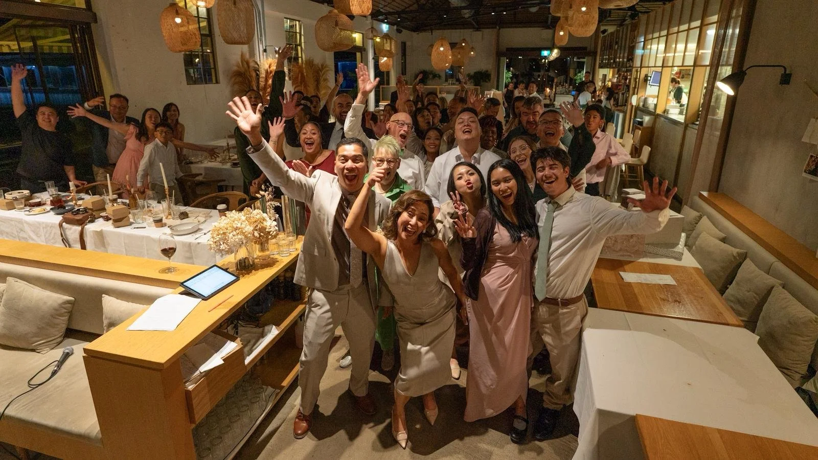 Group of people smiling and waving at a wedding in a restaurant or event space with decorated tables and hanging lights.