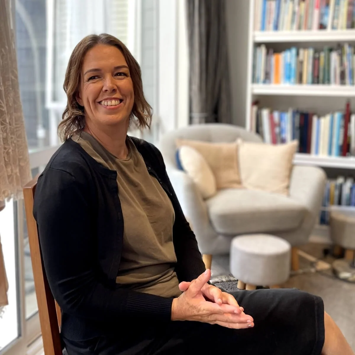 A woman with short brown hair, smiling, sitting in a living room with a beige armchair, bookshelves, and light coming through a window.