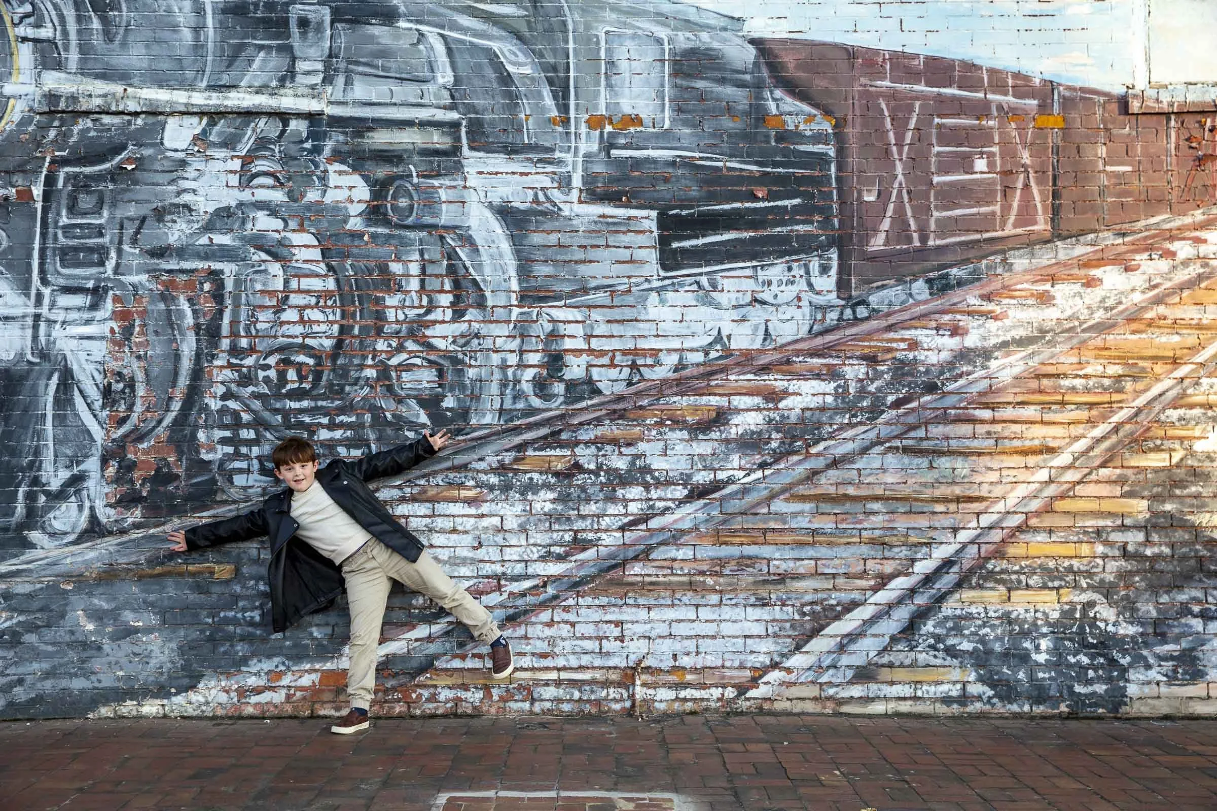 A boy in a black jacket and beige pants standing in front of a mural of a train painted on a brick wall, with his arms outstretched and one foot on the ground.