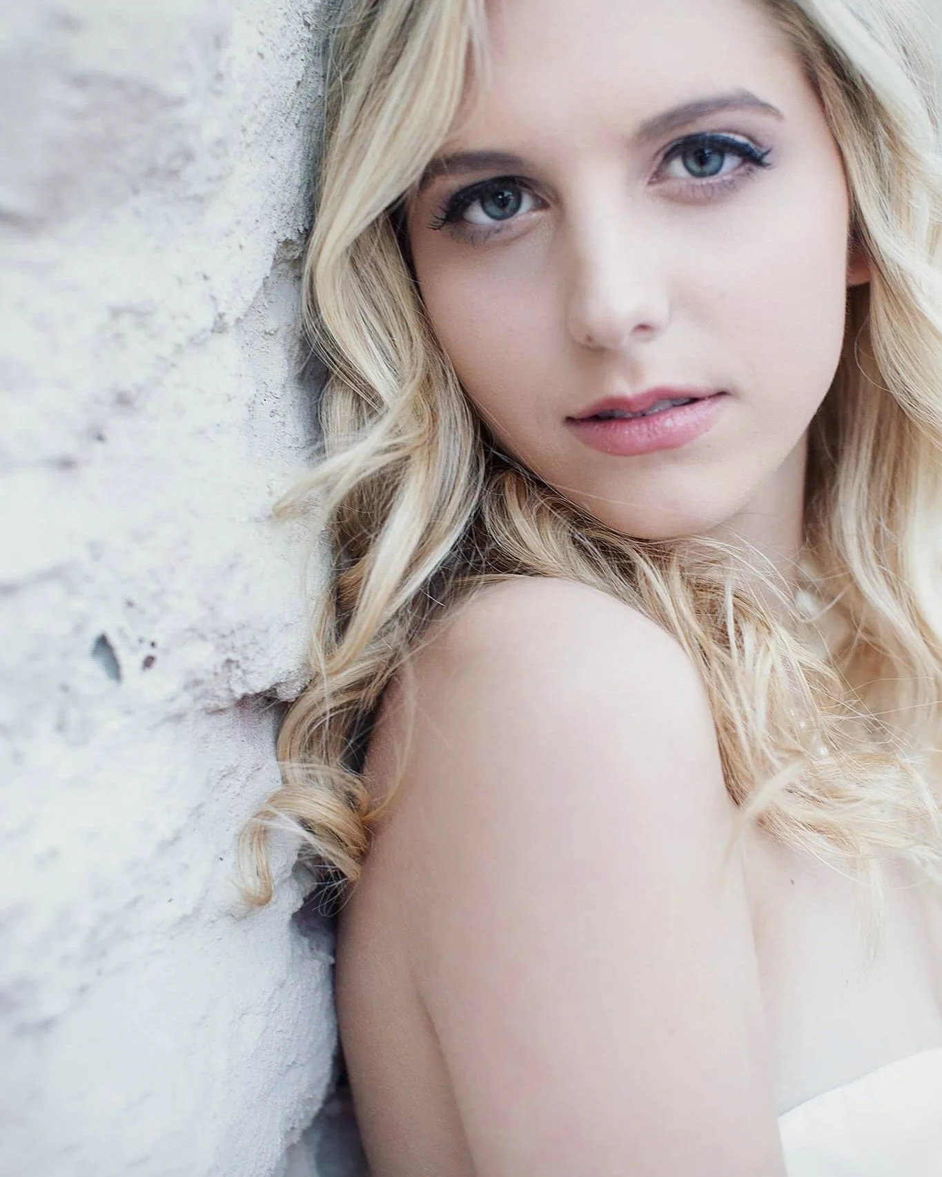 A young woman with blonde hair and blue eyes looks at the camera against a textured white wall.