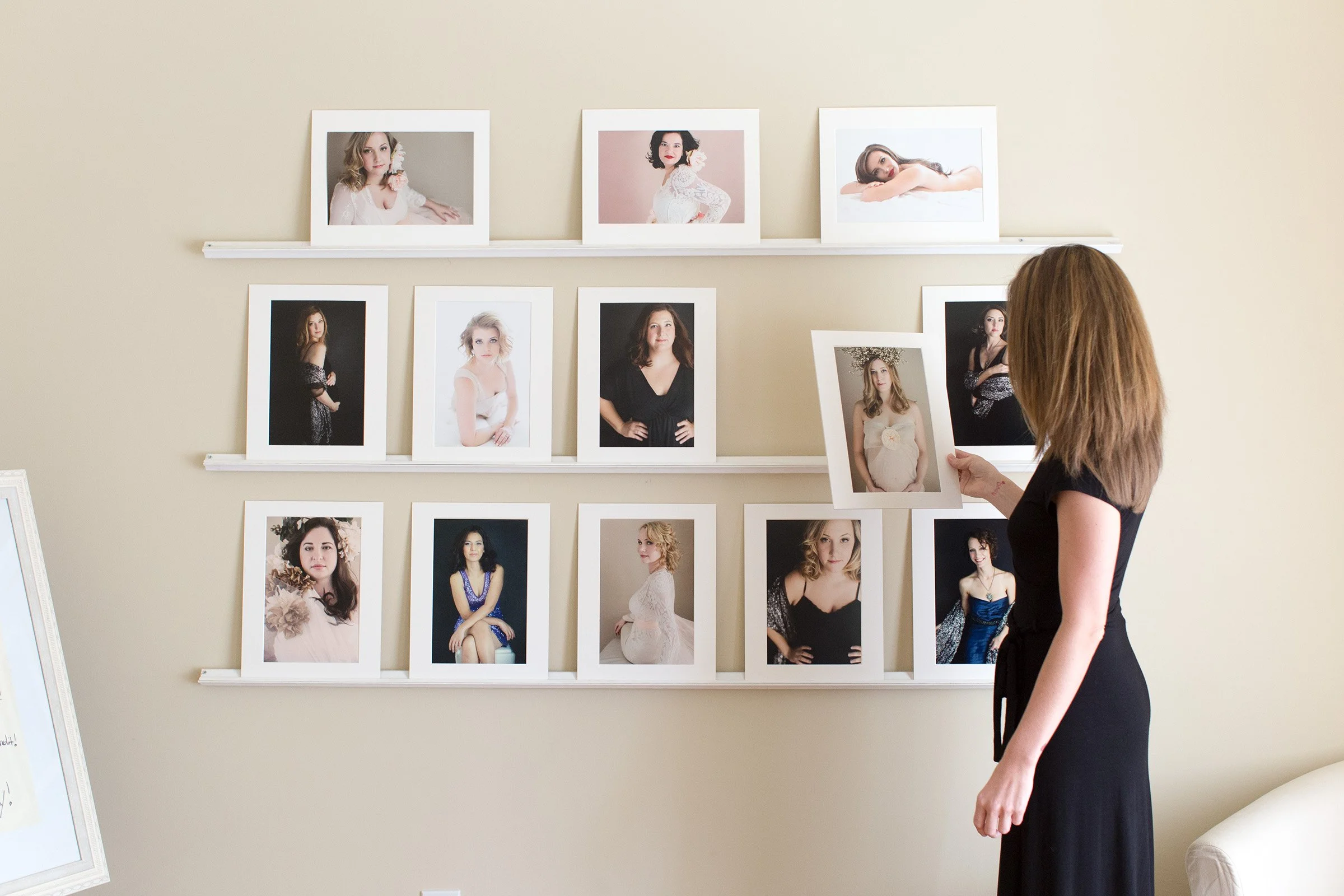 A woman with brown hair in a black dress is looking at a photo of a pregnant woman on a wall with multiple framed photographs of women.