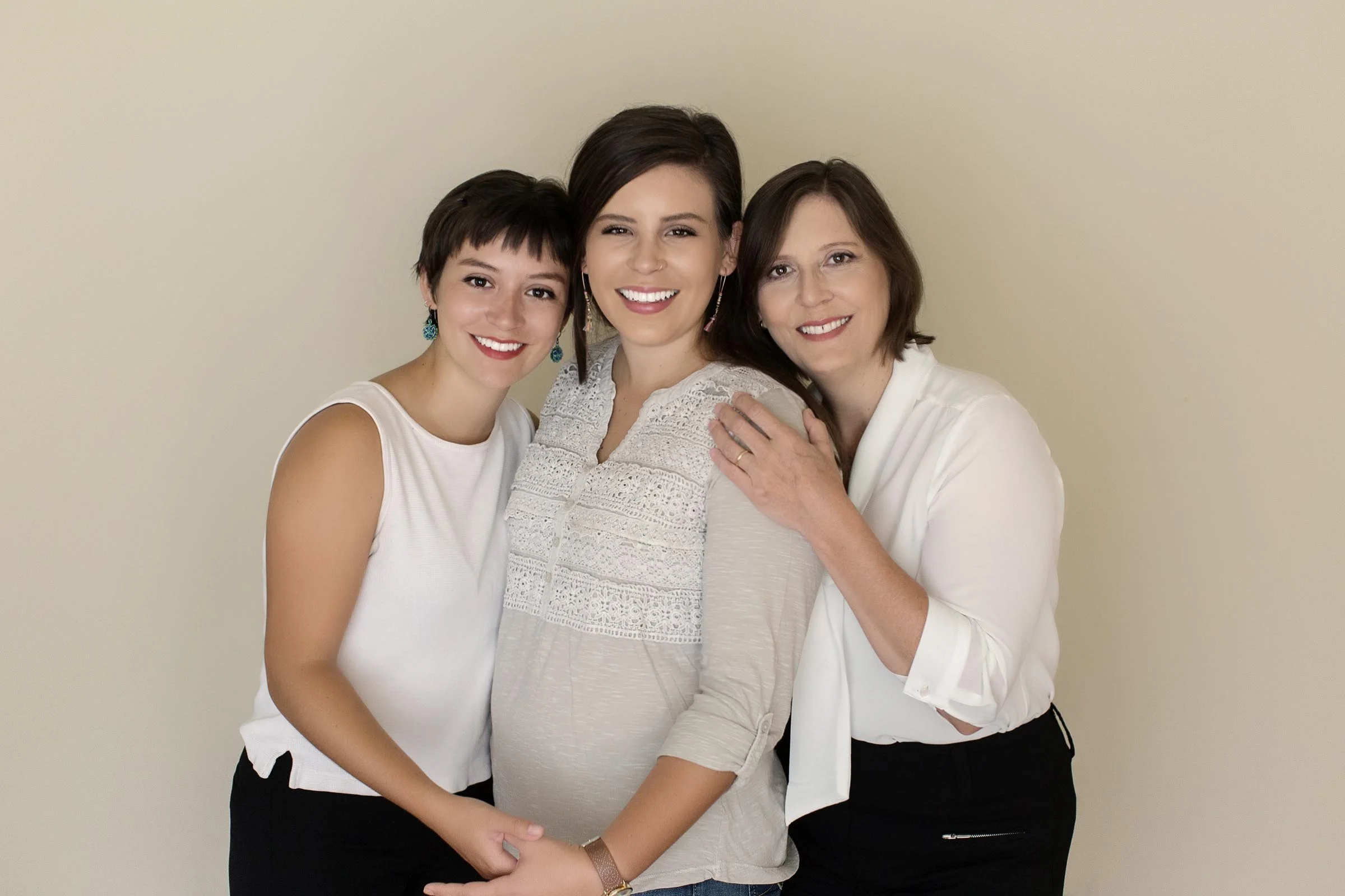 Three women smiling and hugging against a beige background, one pregnant woman in the center, flanked by two women.