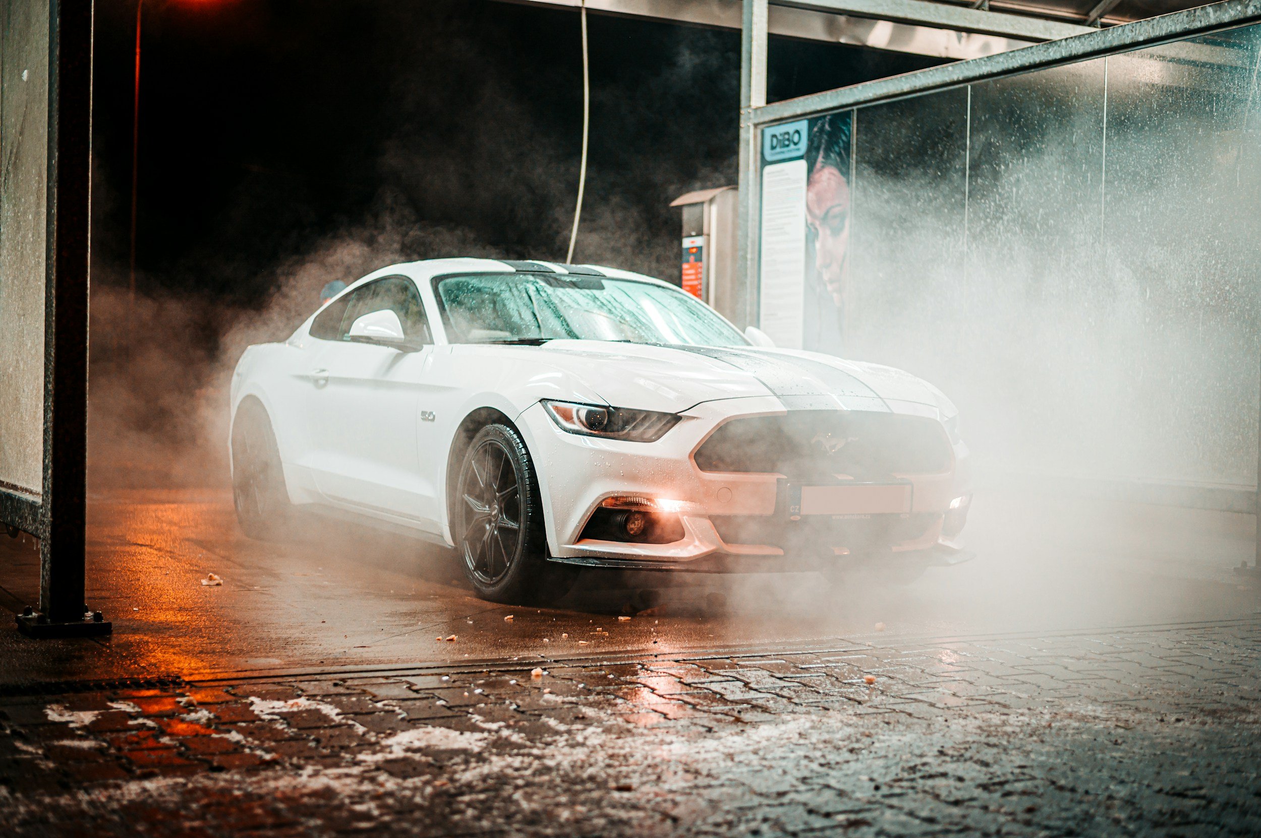 White Ford Mustang being washed at an automated car wash with water and soap, surrounded by mist and foam.