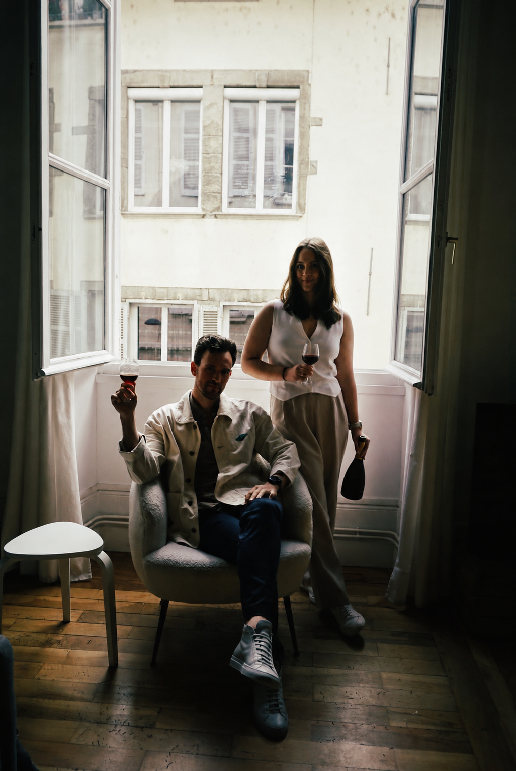 A man and a woman holding glasses of red wine in a room with large open windows, wooden floor, and a view of neighboring buildings.