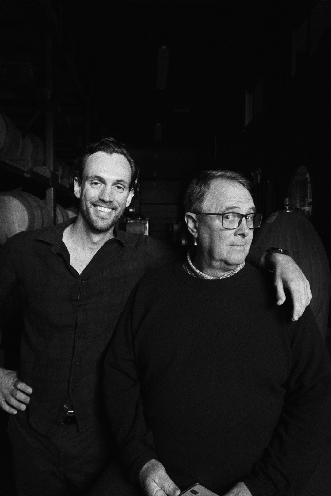 Two men standing together, one with glasses and short hair, the other smiling with longer hair, in a dimly lit room with shelves of barrels in the background.