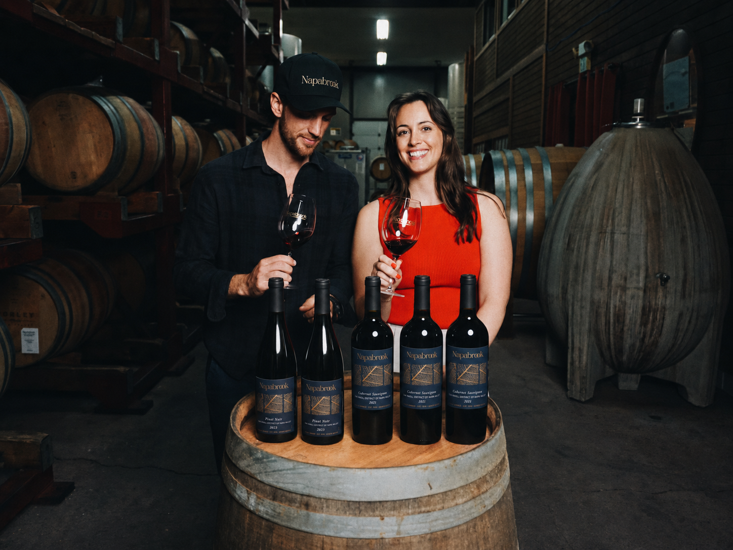 A man and woman standing behind a wine barrel, holding glasses of red wine, with four bottles of wine on the barrel, in a wine cellar with barrels on shelves in the background.