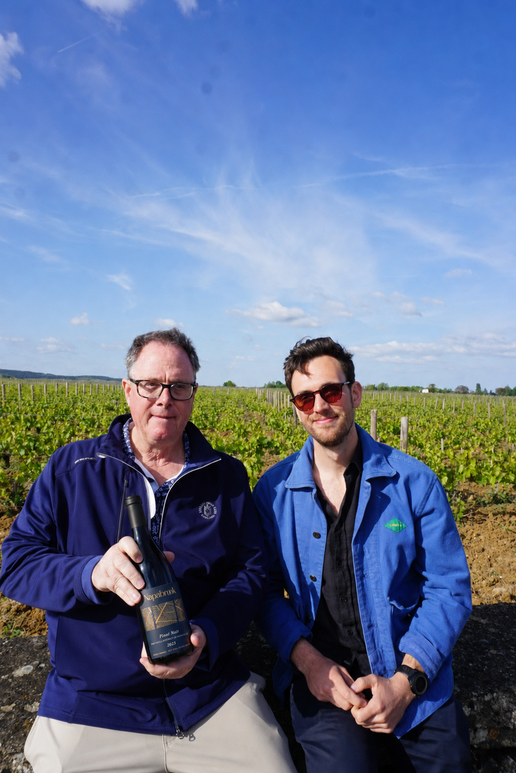 Two men standing in a vineyard, one holding a bottle of wine, under a blue sky with some clouds.