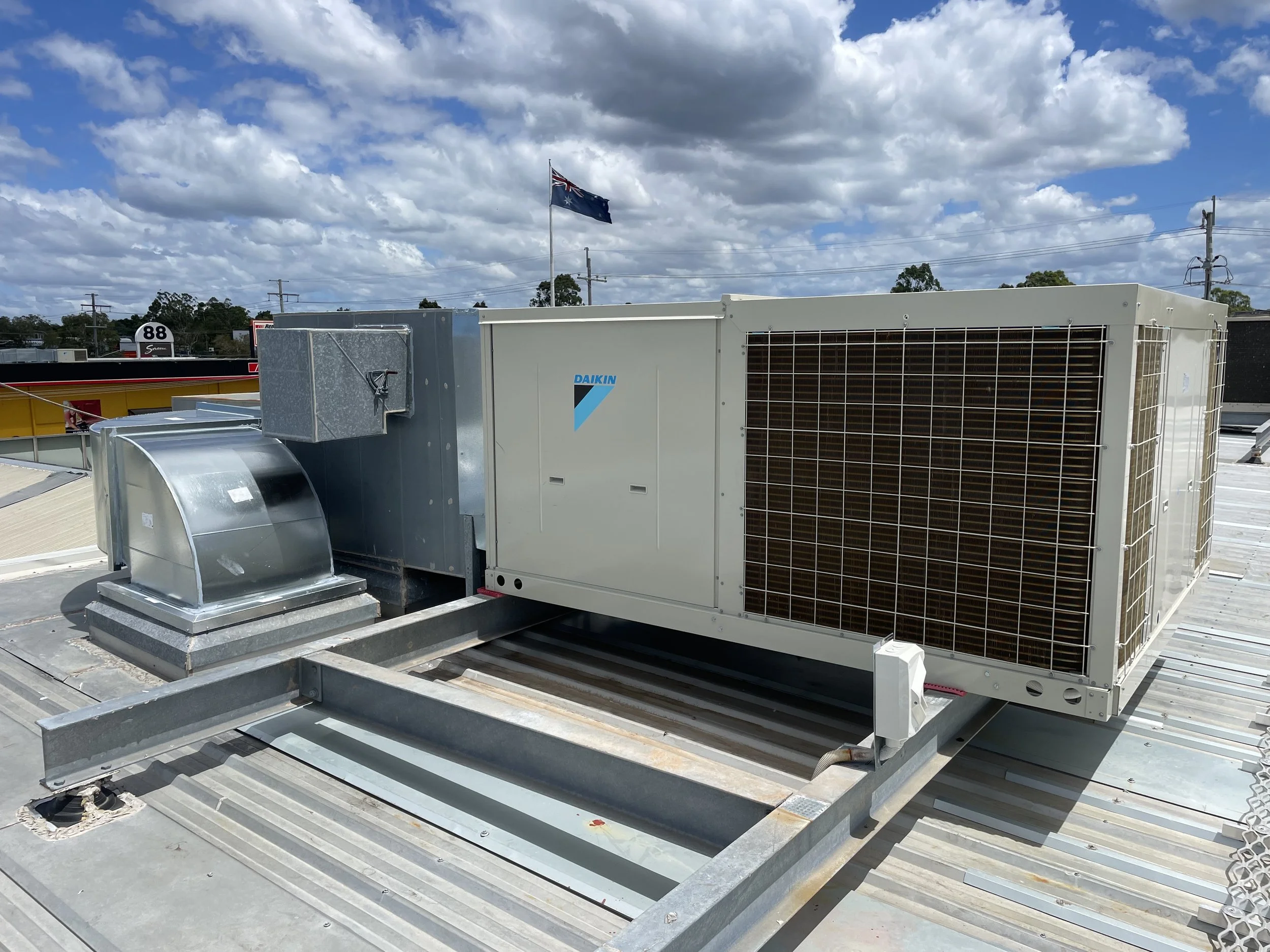 HVAC rooftop unit with the Daikin logo, situated on a metal rooftop with a cloudy sky and an Australian flag in the background.