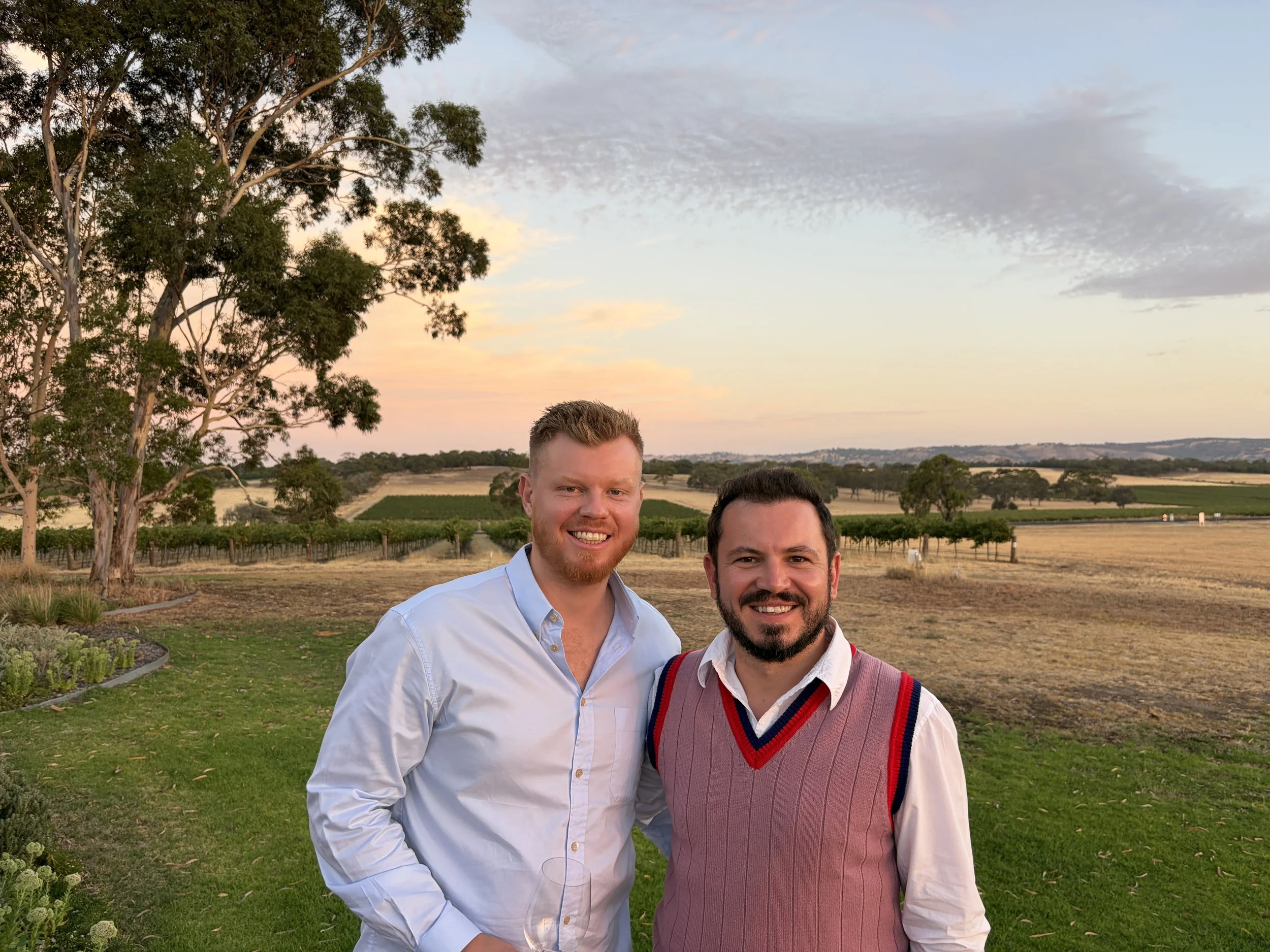 Two smiling men standing outdoors on a grassy area with trees and open fields in the background during sunset.