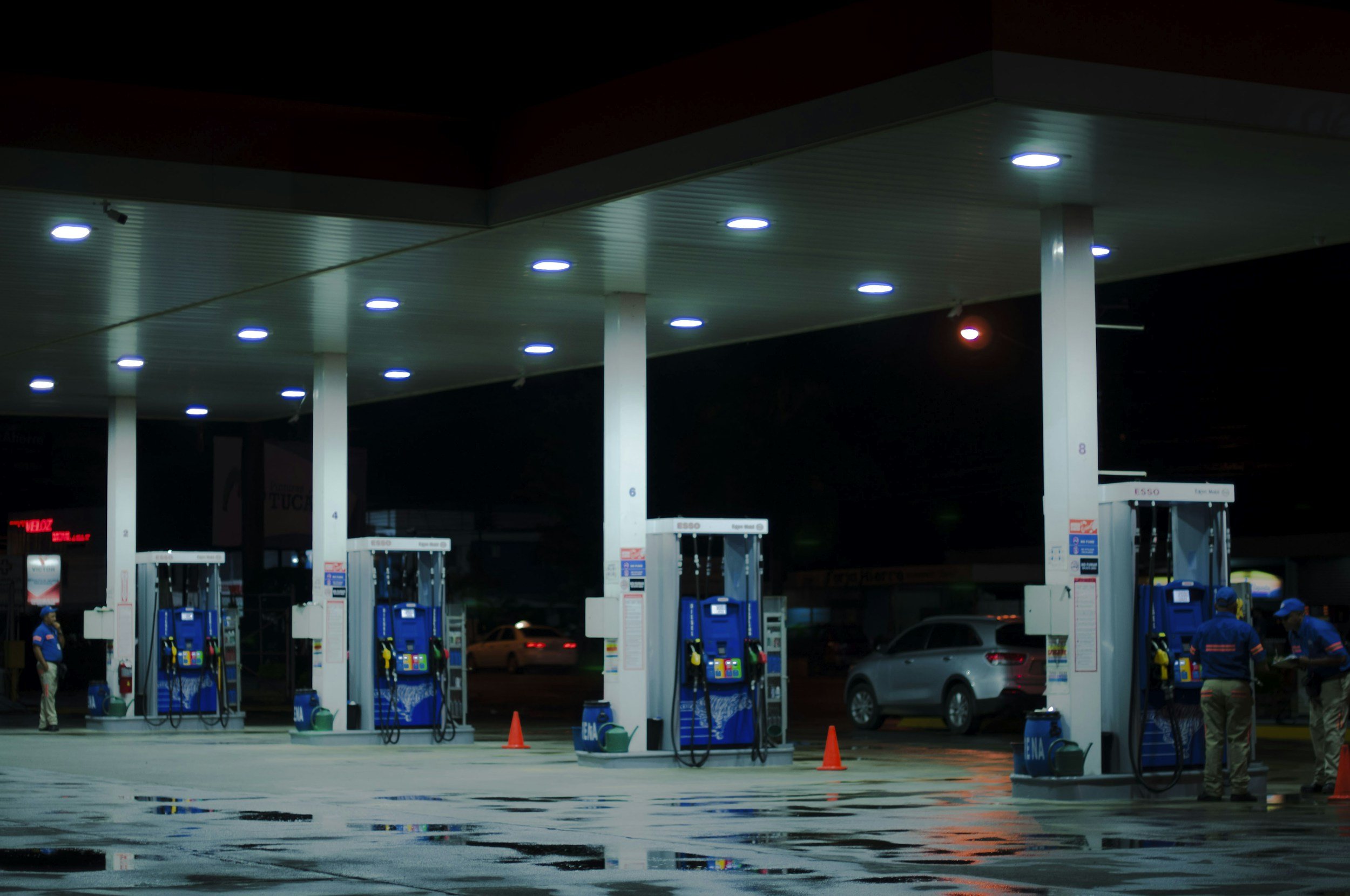 Night scene at a gas station with multiple fuel pumps, illuminated by ceiling lights, with cars and gas station attendants in the background.