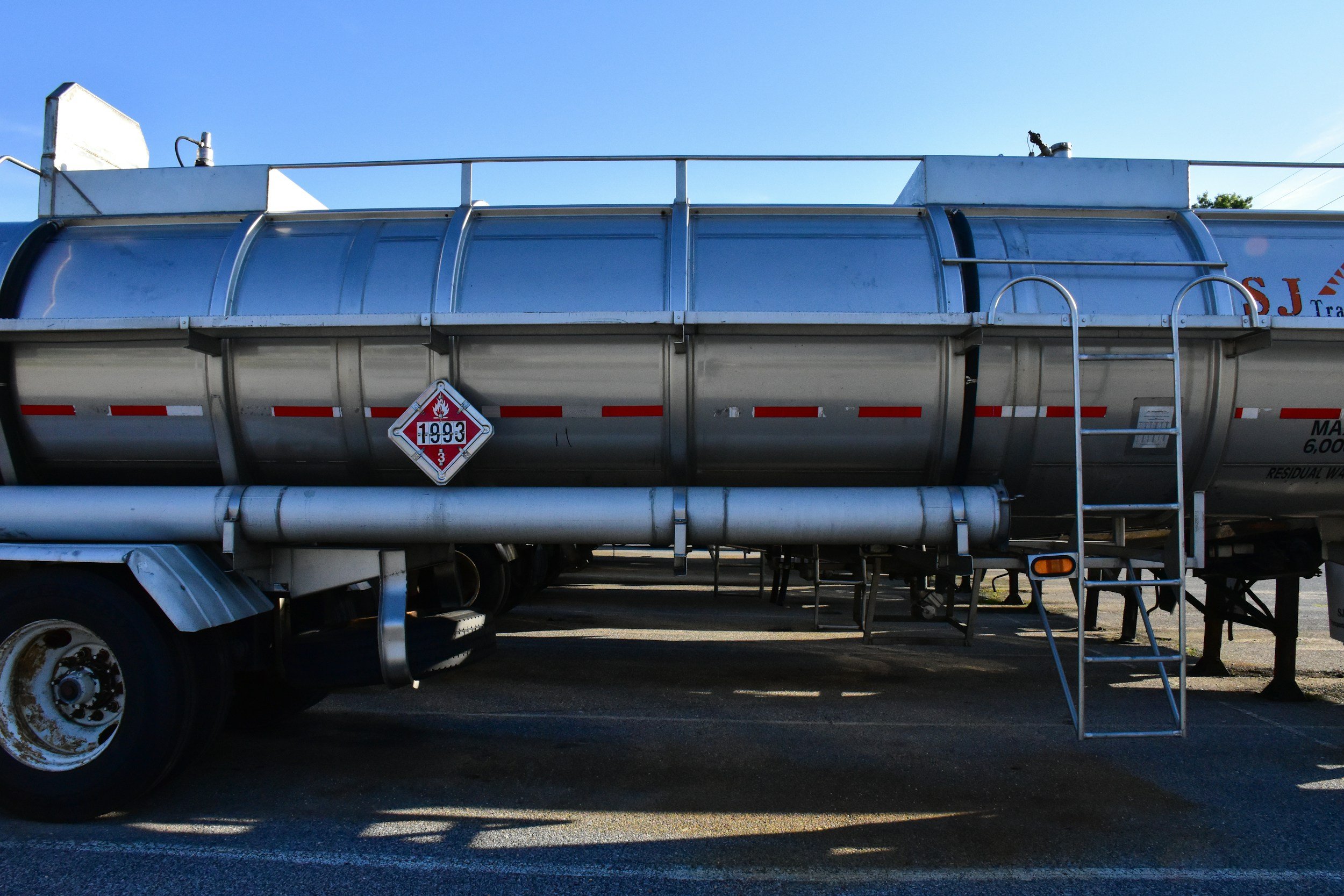 A large silver tanker truck with a hazardous materials warning label and a metal ladder leading to the top, parked on a paved surface under a clear blue sky.