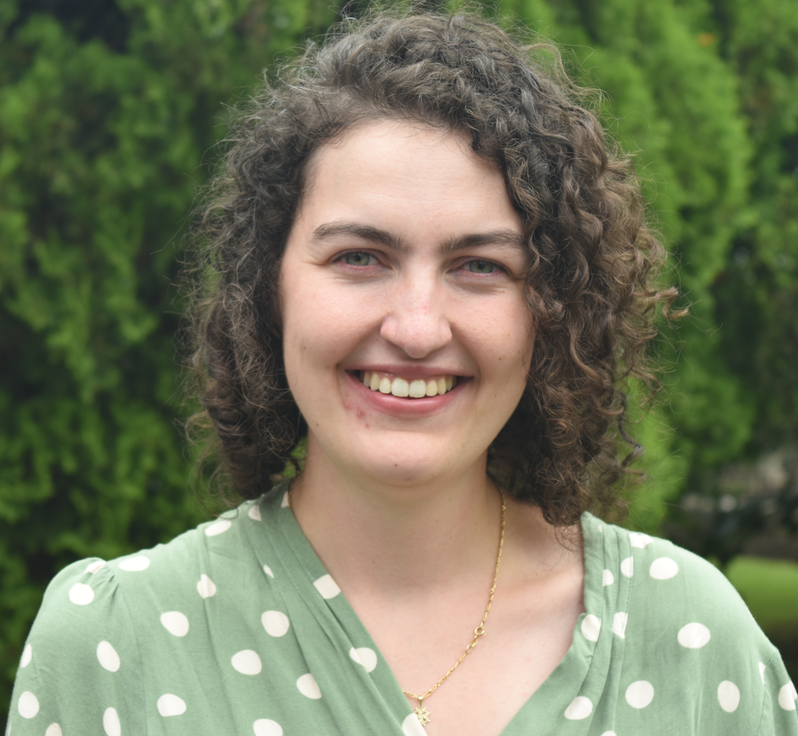 A woman with curly brown hair and light skin smiling outdoors, wearing a green polka-dot dress and gold necklace, with green trees in the background.